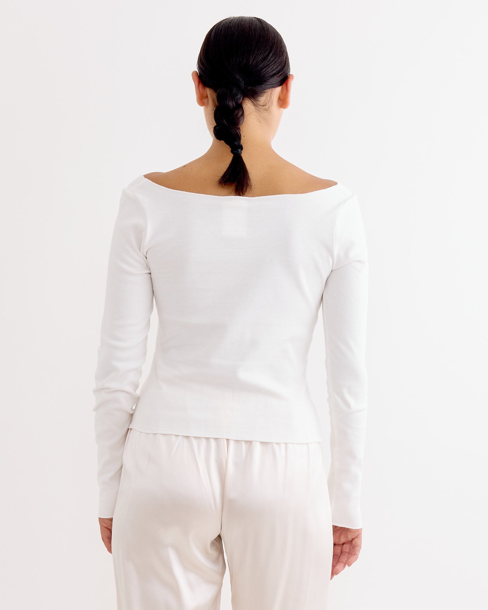 A person with dark braided hair, seen from behind, wears the Hai Gretchen Tee in White—an organic cotton jersey long-sleeve v-neck—paired with light pants against a plain white background.