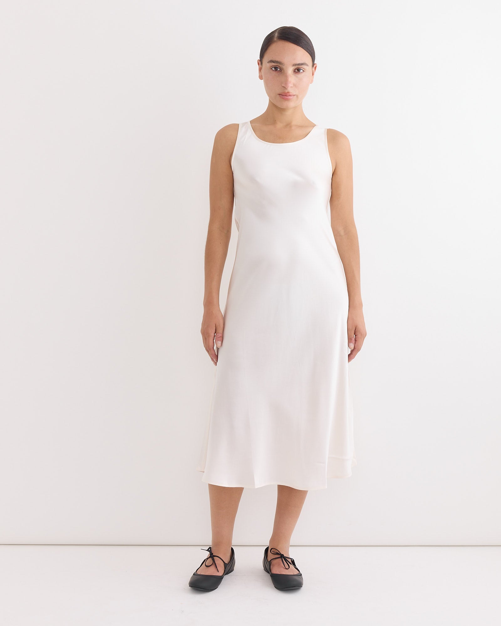 A woman with dark hair pulled back stands against a white background wearing the Hai Roma Dress in Ivory and black ballet flats. She faces forward with a neutral expression.