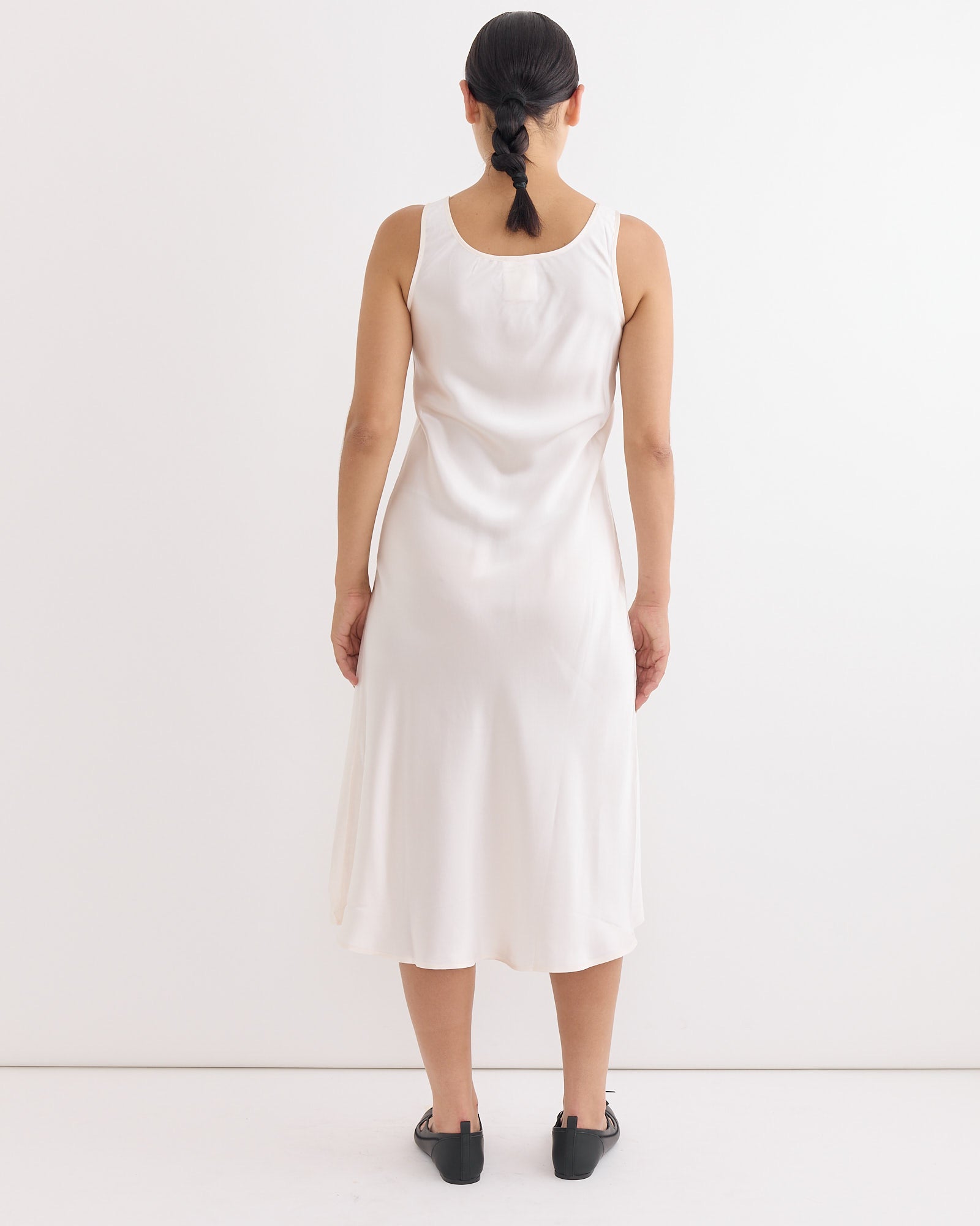 A woman with dark hair in a low braid, wearing Hai's Roma Dress in Ivory and black shoes, stands facing away from the camera against a plain white background.