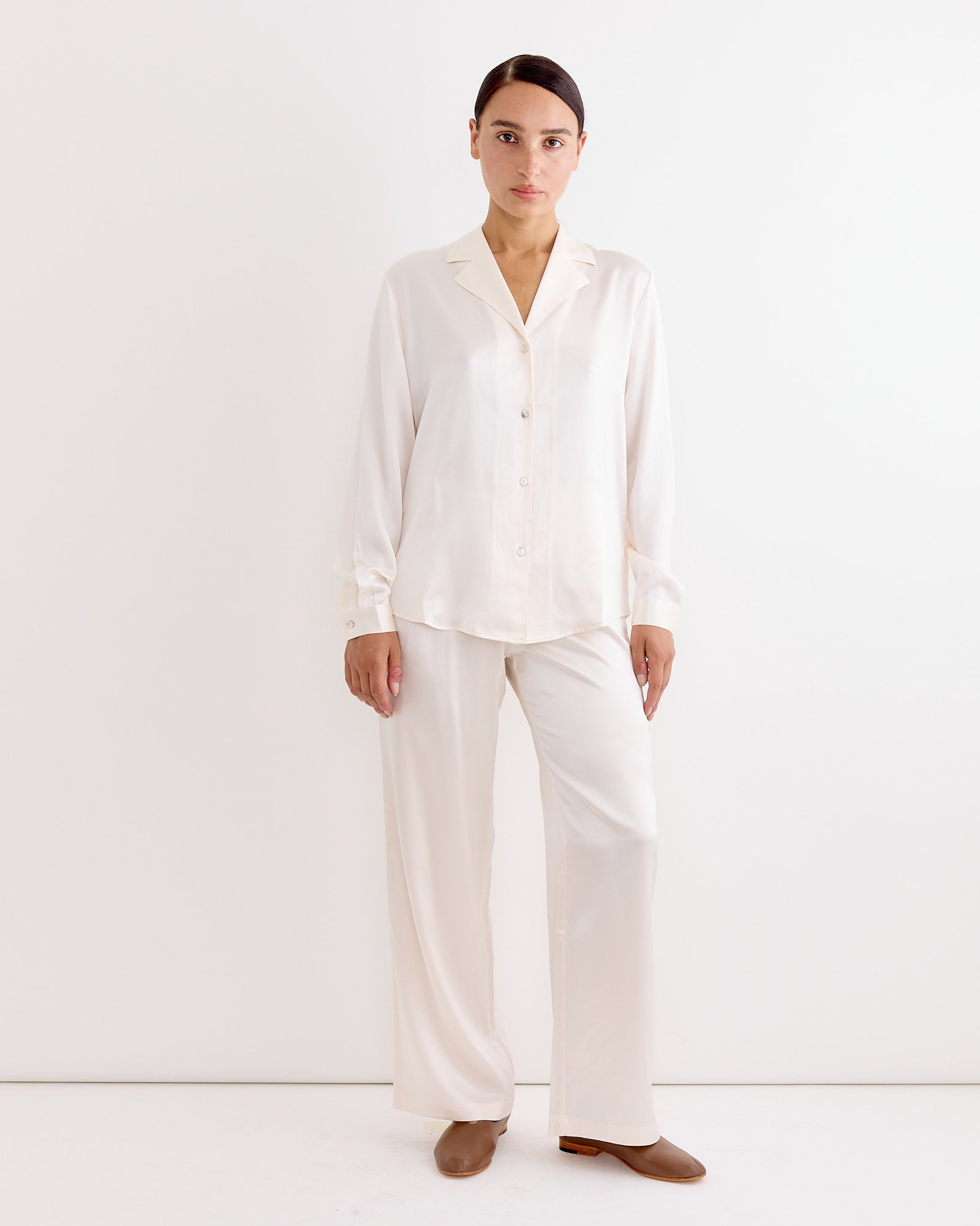 A woman stands against a plain white background, wearing the Hai Soma Trouser in Ivory with a matching long-sleeve shirt and brown shoes. She faces forward with a neutral expression.