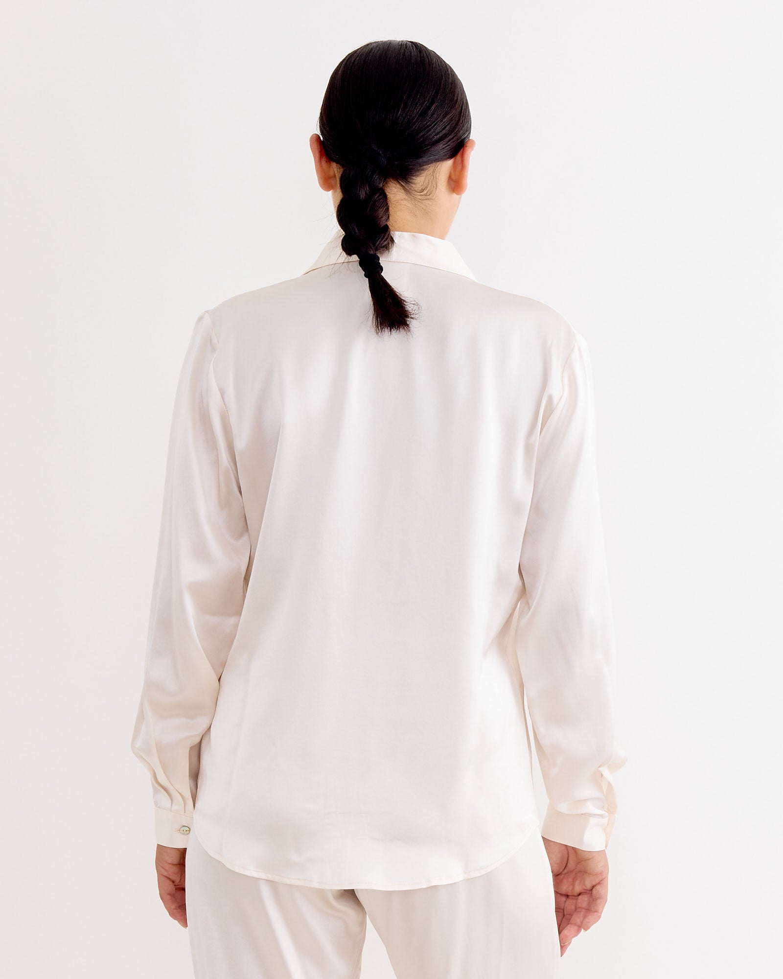 A person with dark hair in a low braid is seen from behind, wearing the Hai Irene Blouse in Ivory paired with matching pants, set against a plain white background.