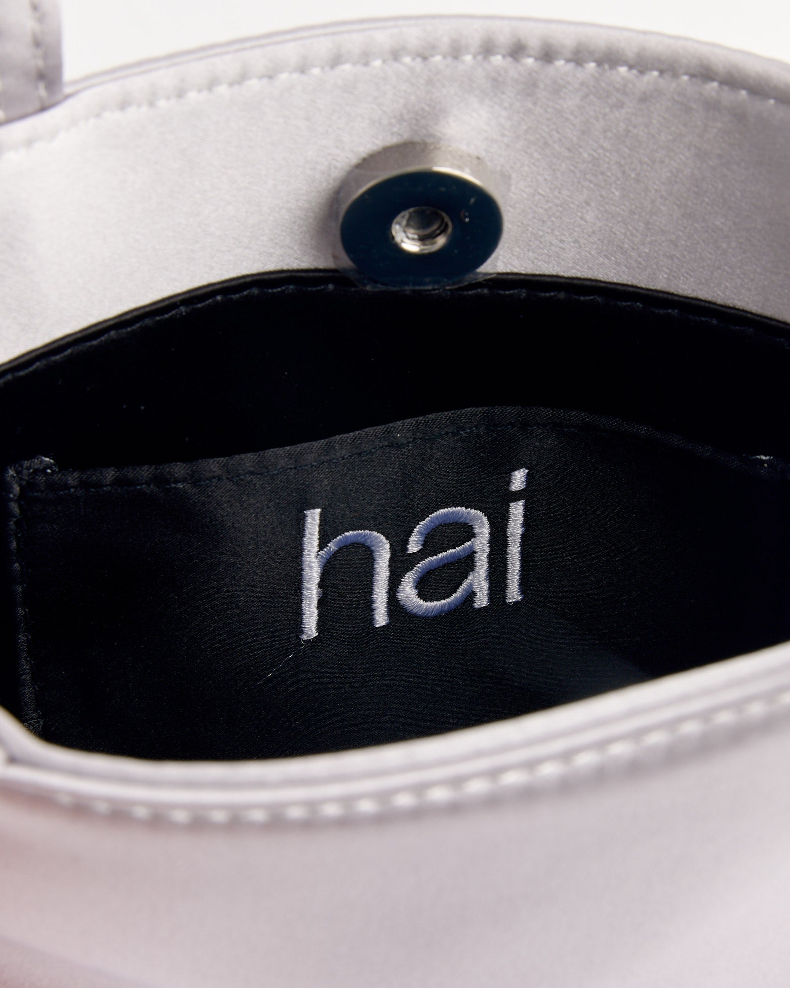 Close-up of the Little Silk Bag in Silver by Hai, showing the silver satin interior, snap button, and black inner label with the embroidered "hai" logo, emphasizing its chic design.