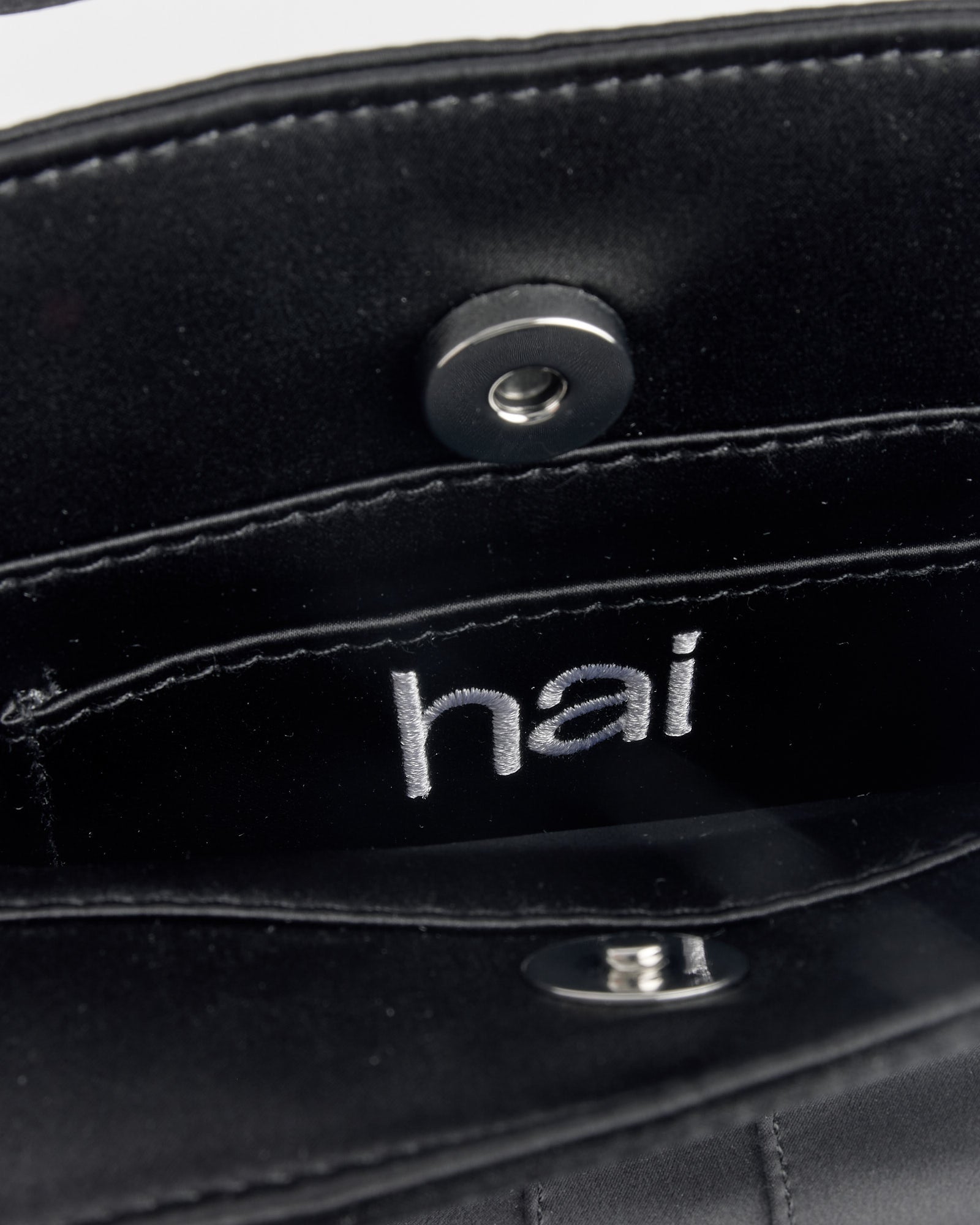 Close-up of the inside of the Hai Farfalle Bag in Black, showing "hai" embroidered in white on the inner pocket and a visible snap button closure.