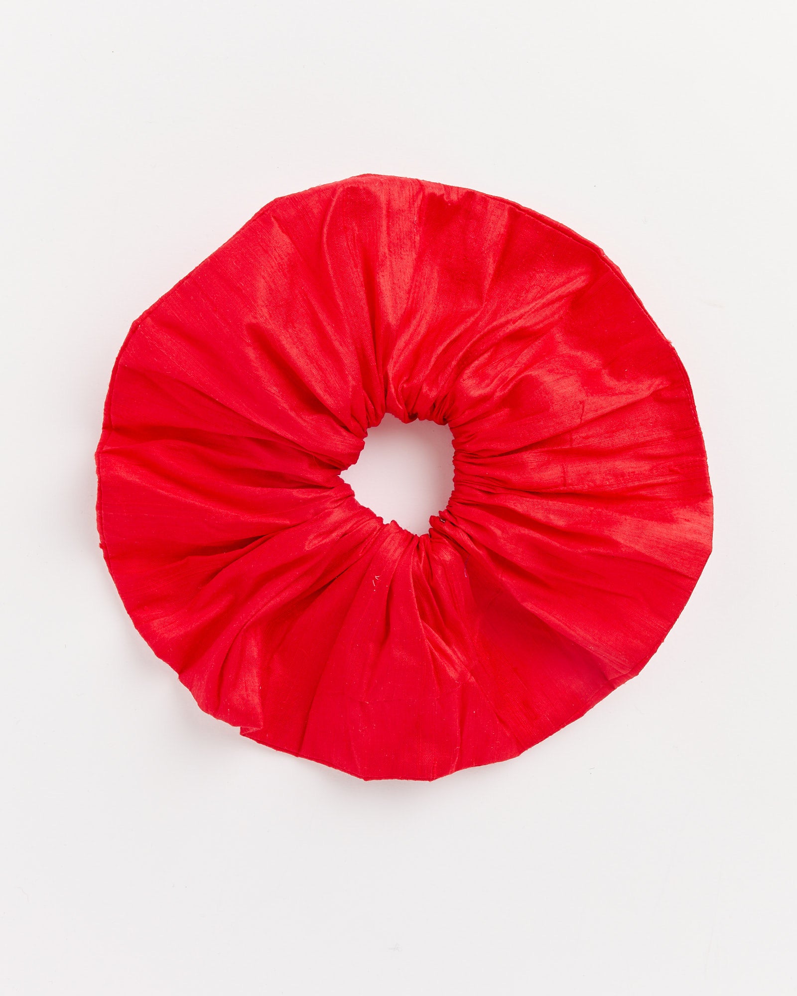 A vibrant red Silk Scrunchie by Hai is shown against a white background, featuring circular gathered shiny silk with a visible elastic center.