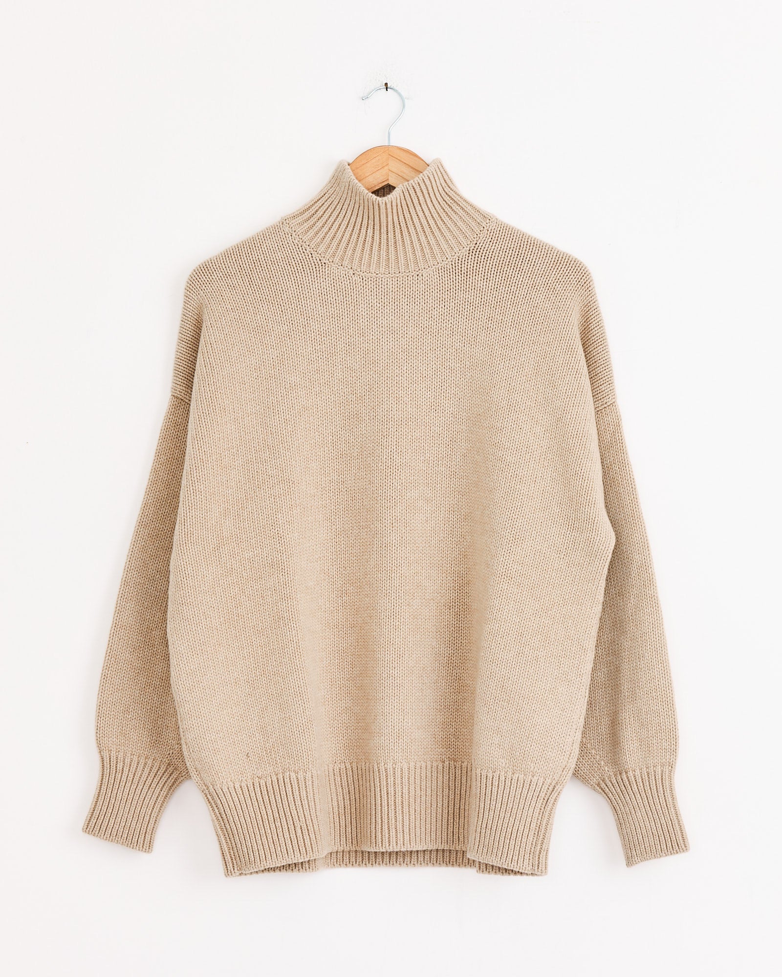 The Cordera Merino Wool Chunky Turtleneck in Beige, featuring long sleeves, a ribbed texture, and an oversized fit, is displayed on a wooden hanger against a plain white background.
