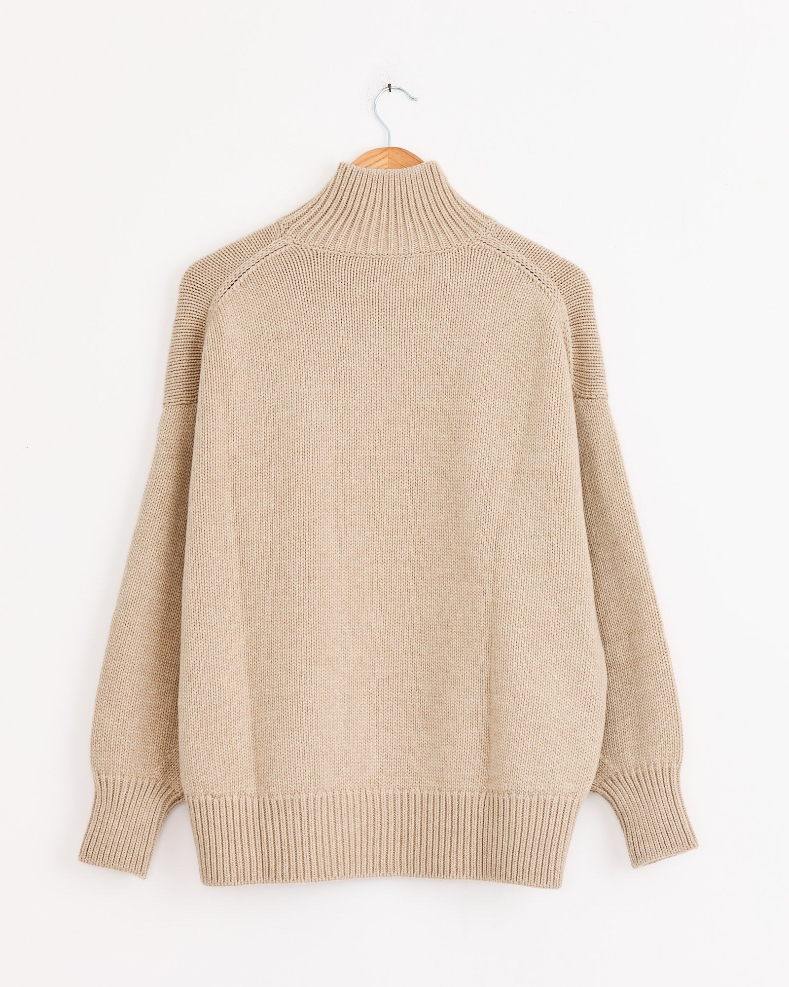 The CORDERA Merino Wool Chunky Turtleneck in Beige, featuring an oversized fit with ribbed cuffs and hem, hangs on a wooden hanger against a plain white wall.
