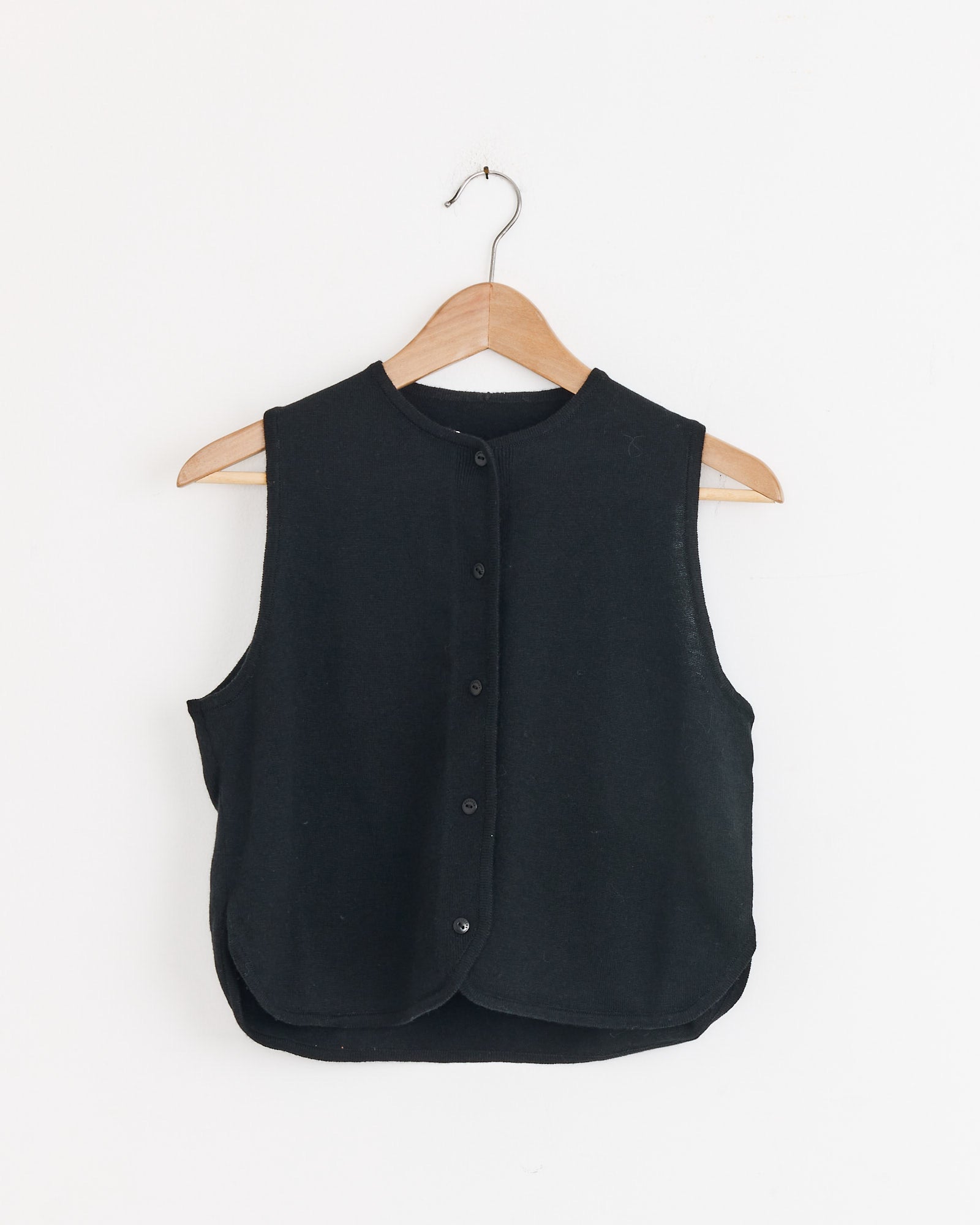 The Cordera Merino Wool Waistcoat in Black is an ethically made, cropped vest with front buttons, shown on a wooden hanger against a white wall.
