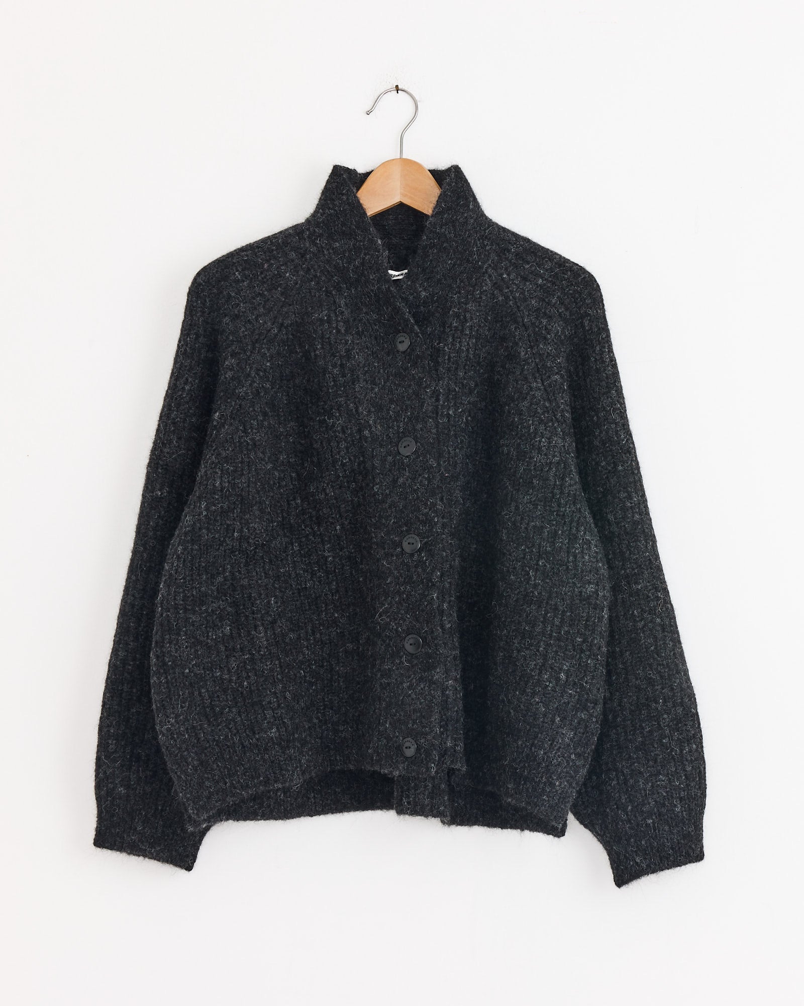 The Cordera Baby Alpaca Cardigan in Anthracite, an oversized, button-up knit with a high collar made in Spain, hangs on a wooden hanger against a plain white wall.