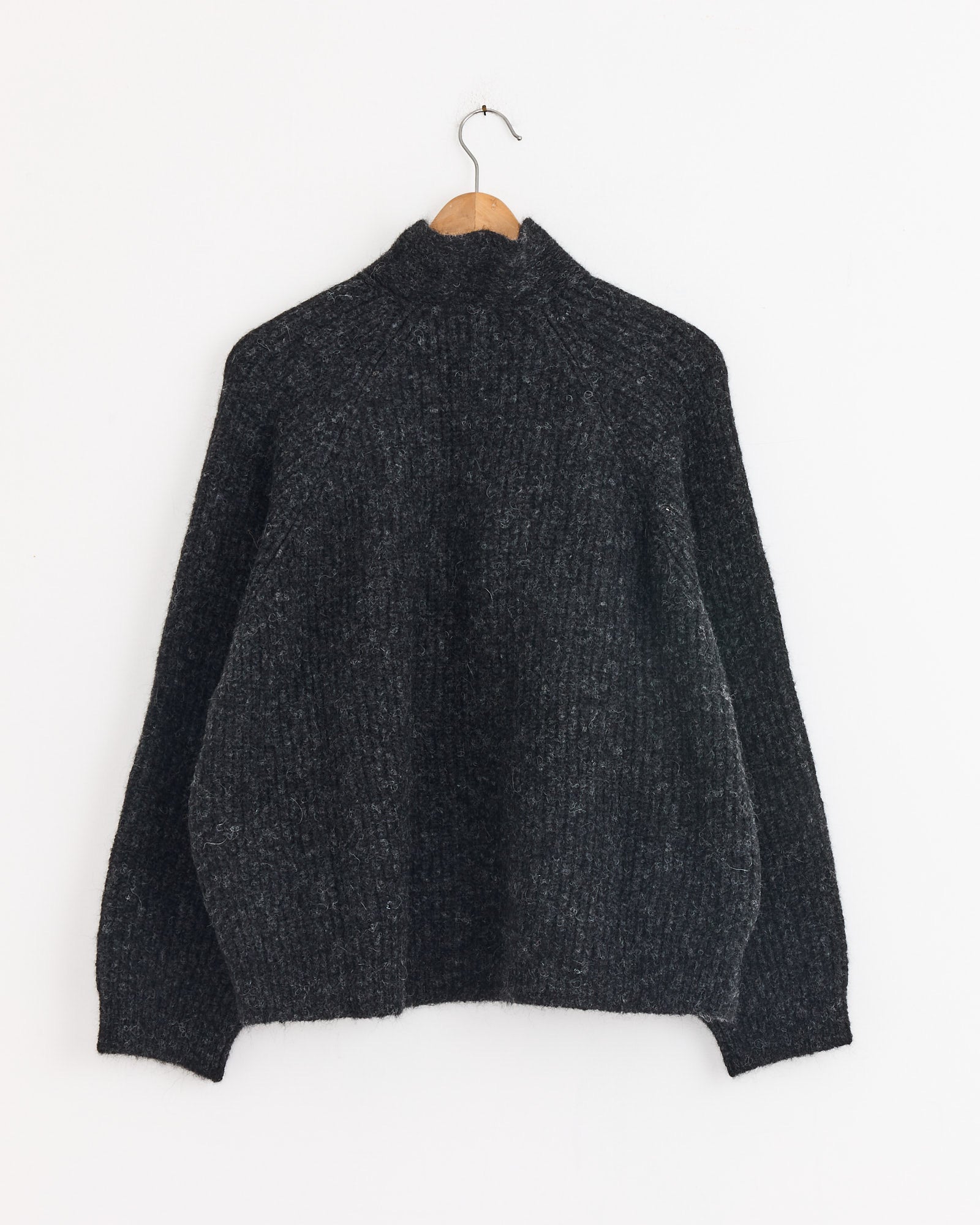 The Cordera Baby Alpaca Cardigan in Anthracite, featuring long sleeves and a high neck, hangs on a wooden hanger against a plain white wall.