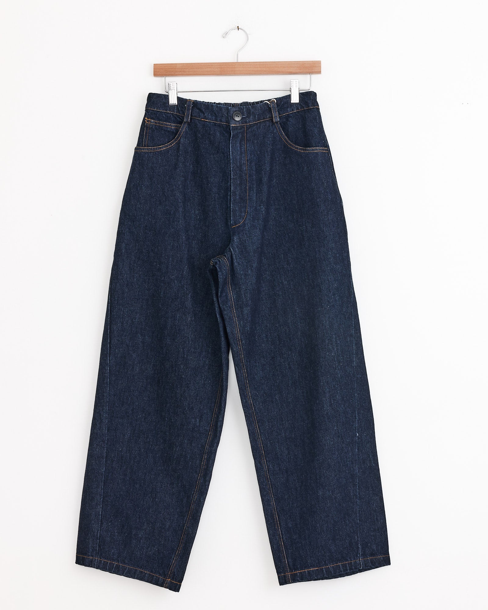 The Cordera Denim Straight Pant in Indigo, featuring a high-rise, wide-leg fit with button and zipper closure and front pockets, hangs on a wooden hanger against a white background. Made in Spain.