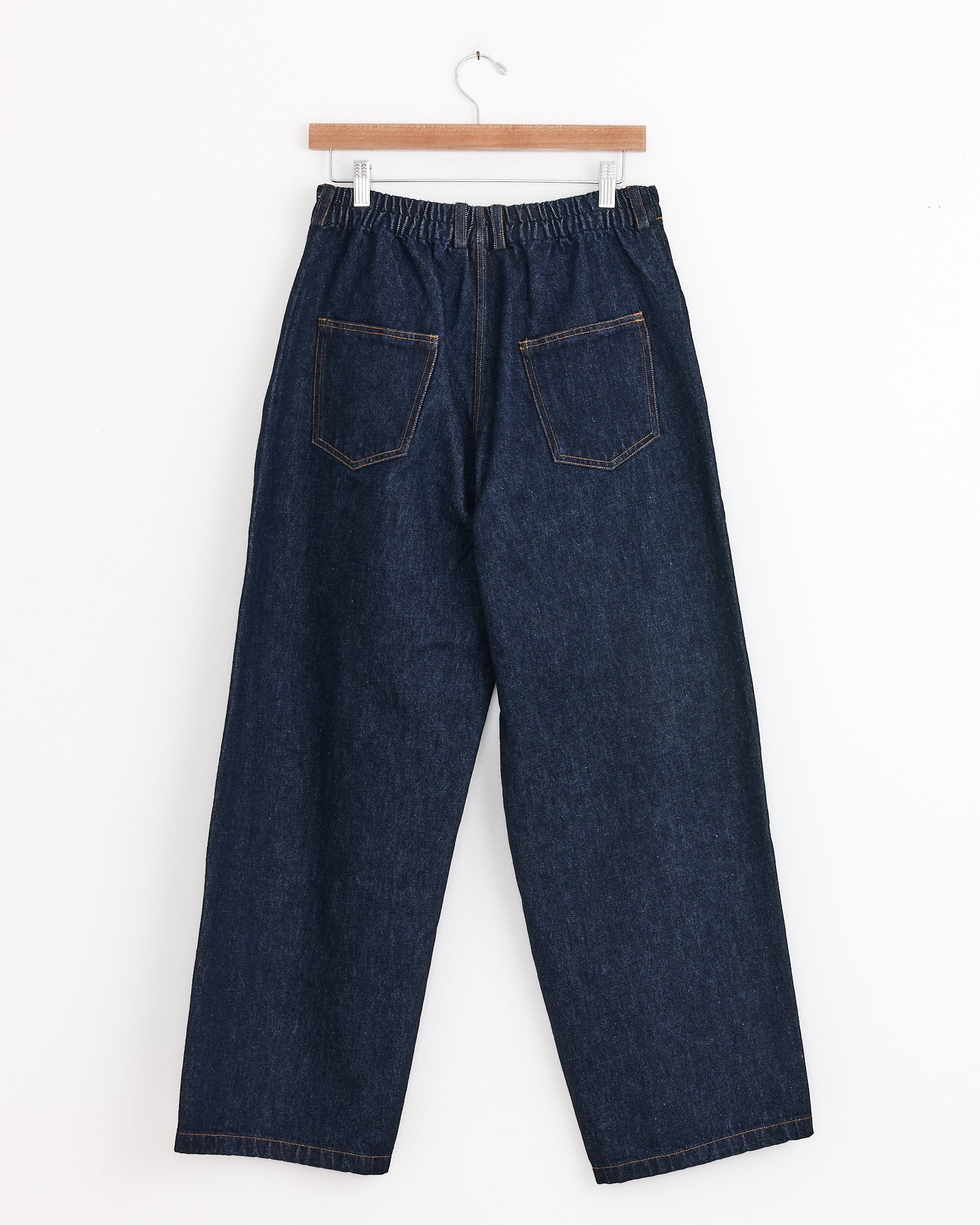 The CORDERA Denim Straight Pant in Indigo features a loose fit, high rise, elastic waistband, and two back pockets, shown hanging on a wooden hanger against a white background.
