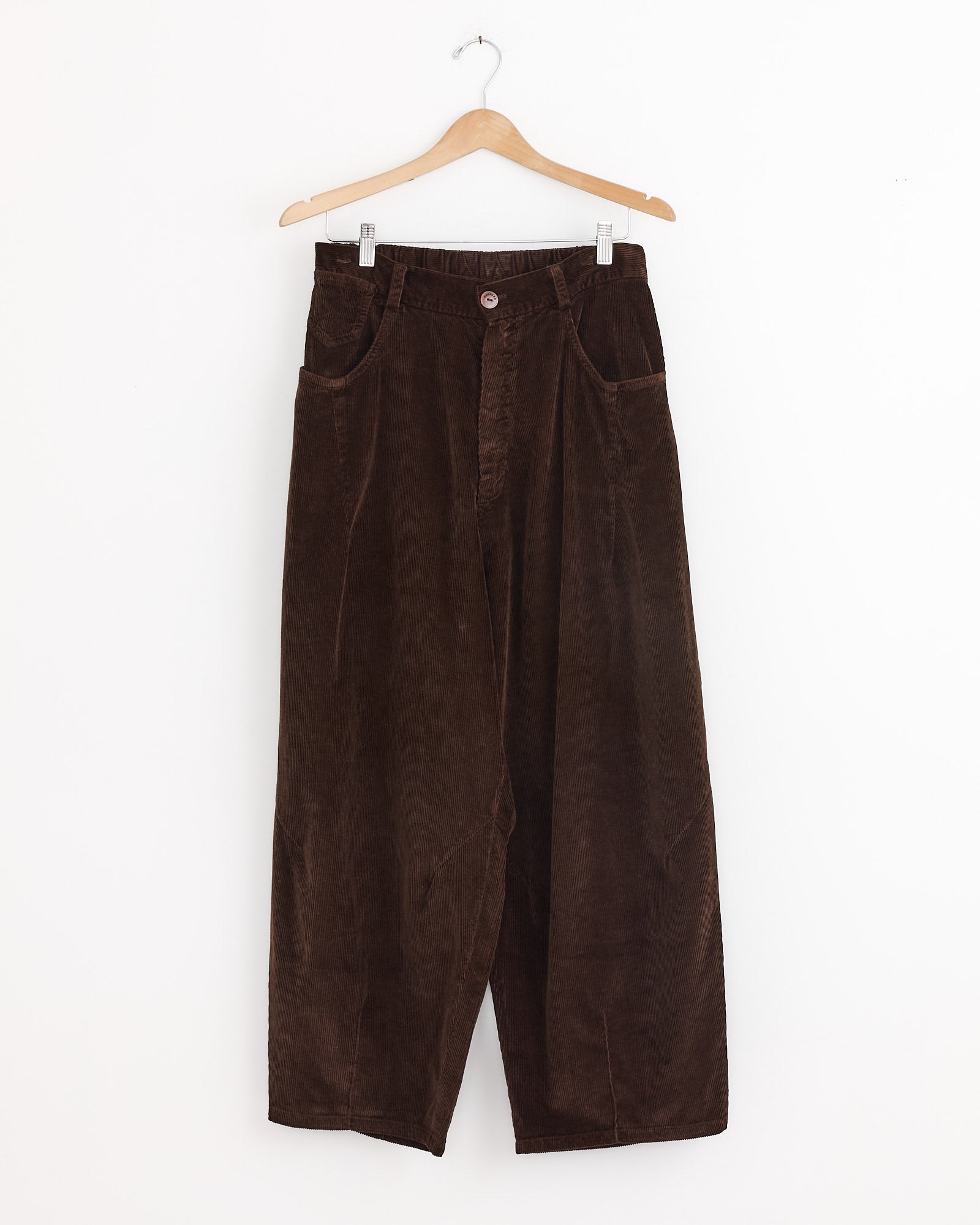 The Cordera Baggy Pant in Brown—a high-waisted, relaxed-fit, wide-leg corduroy pant made in Spain—hangs on a wooden hanger against a plain white background.