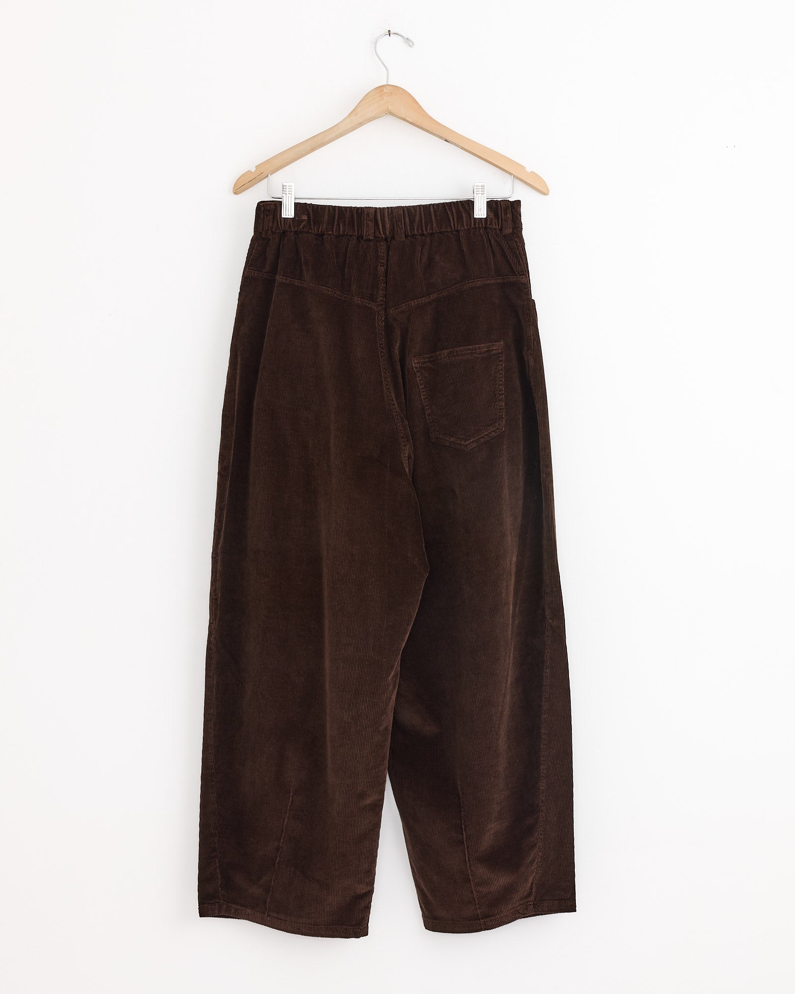 The Cordera Baggy Pant in Brown, made in Spain, features a loose fit, elastic waistband, and single back pocket, shown hanging on a wooden hanger against a white background.