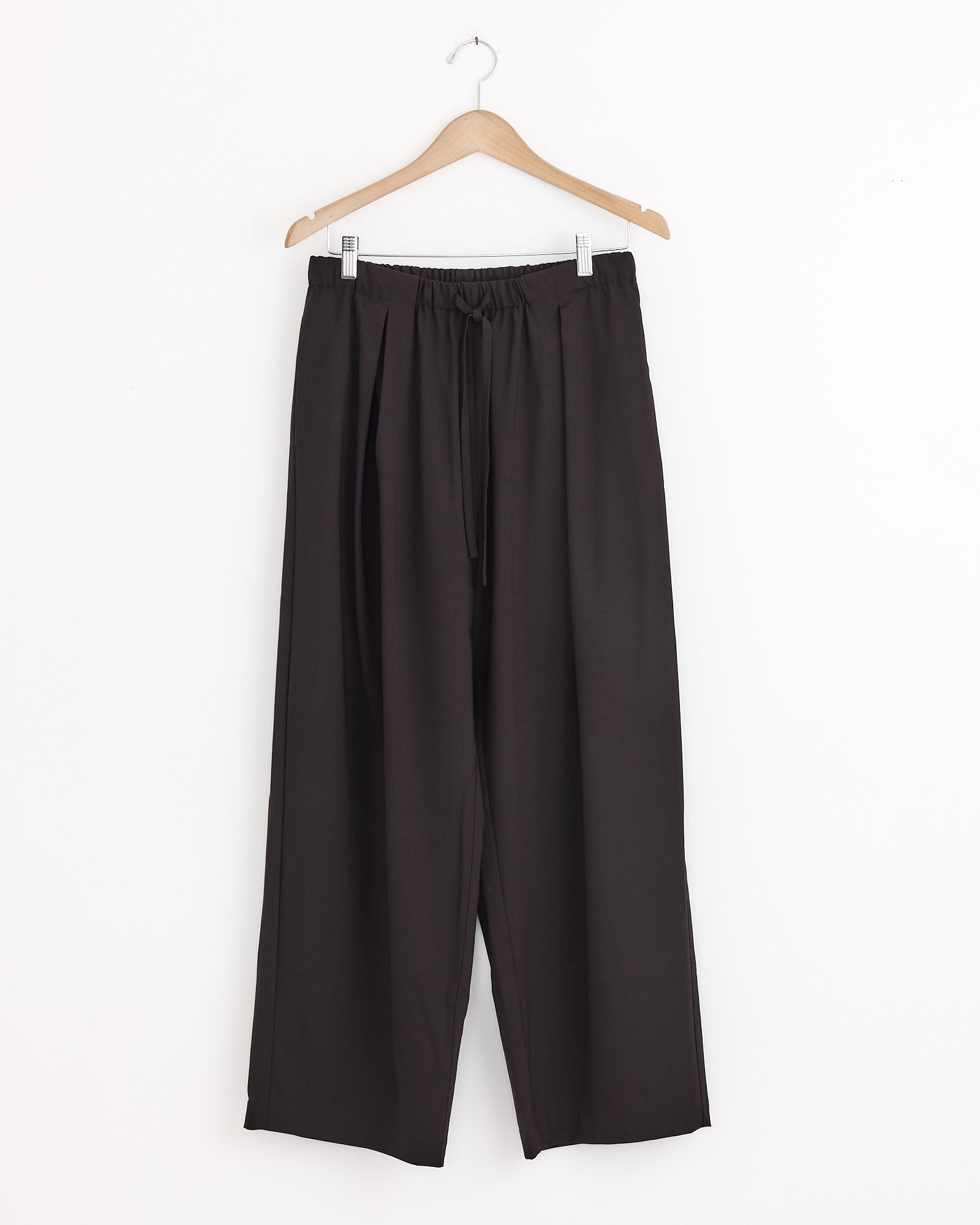 The Cordera Relaxed Pleated Pant in Dark Brown features an elastic waistband and drawstring, crafted from lightweight wool, and hangs on a wooden hanger against a plain white wall.
