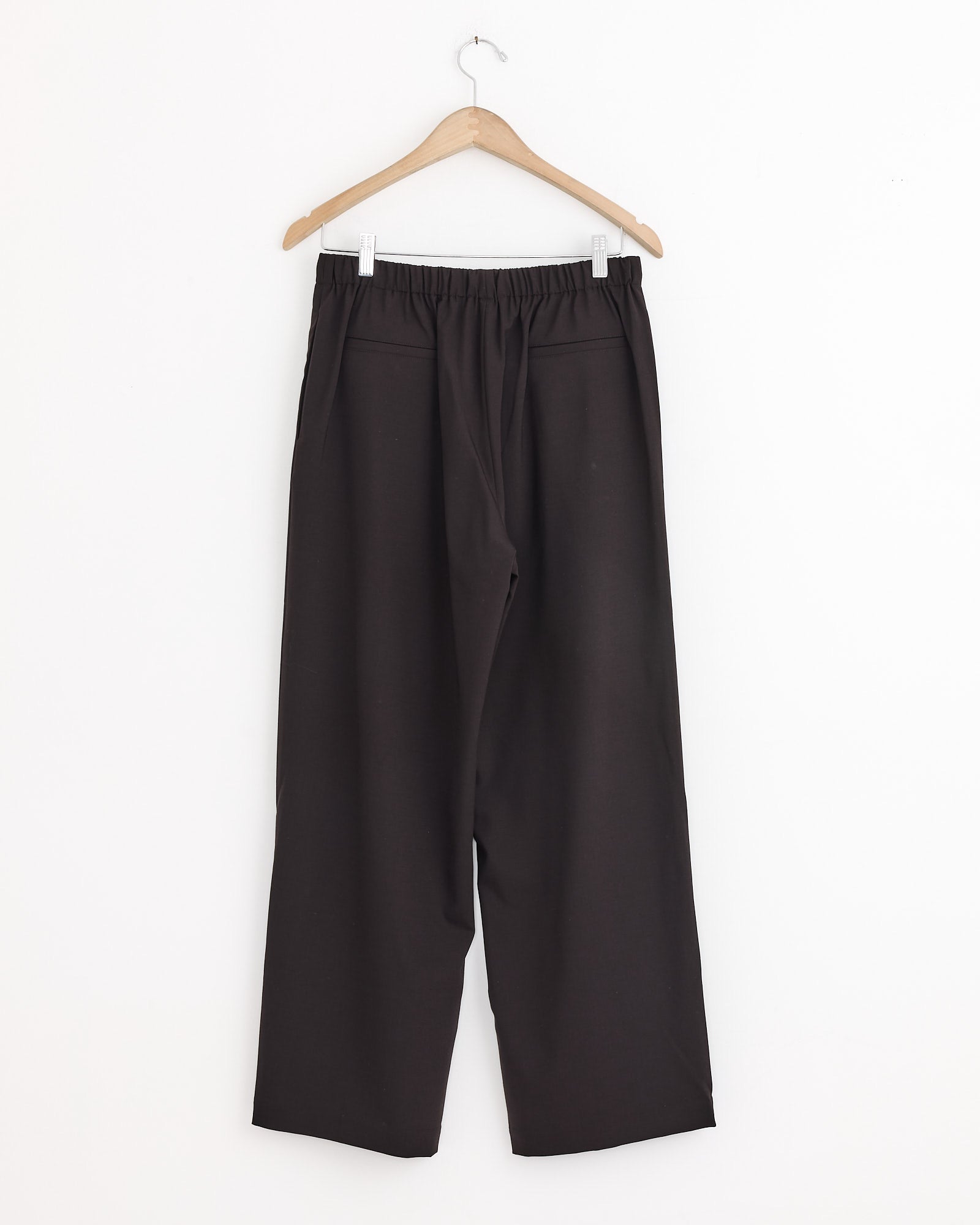 The Cordera Relaxed Pleated Pant in Dark Brown, featuring an elastic waistband and lightweight wool fabric, hangs on a wooden hanger against a plain white wall.