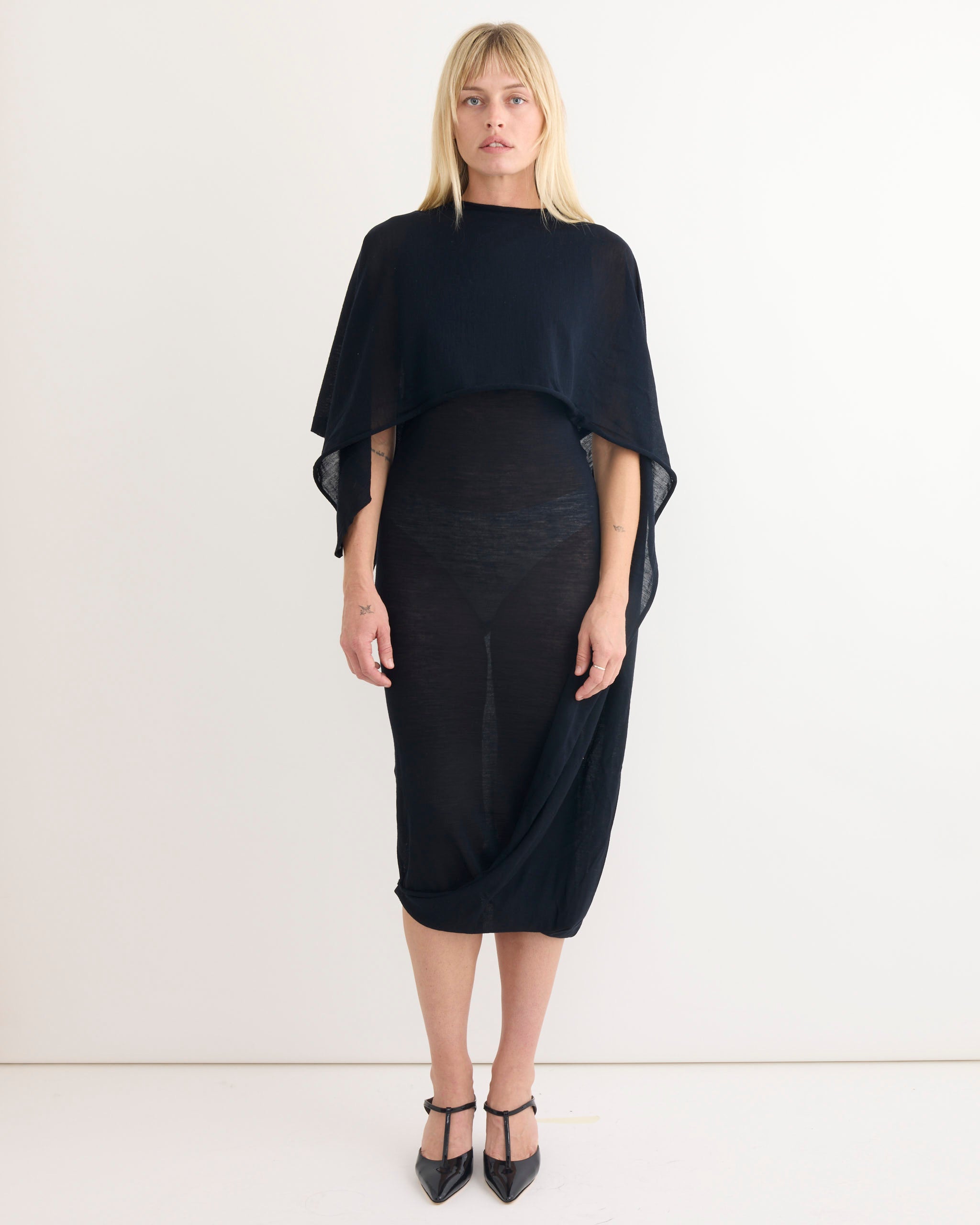 A woman stands against a plain white background, wearing the Paloma Wool Willy Dress in Navy with a draped cape-like top and pointed black heels. She looks straight ahead with a neutral expression.