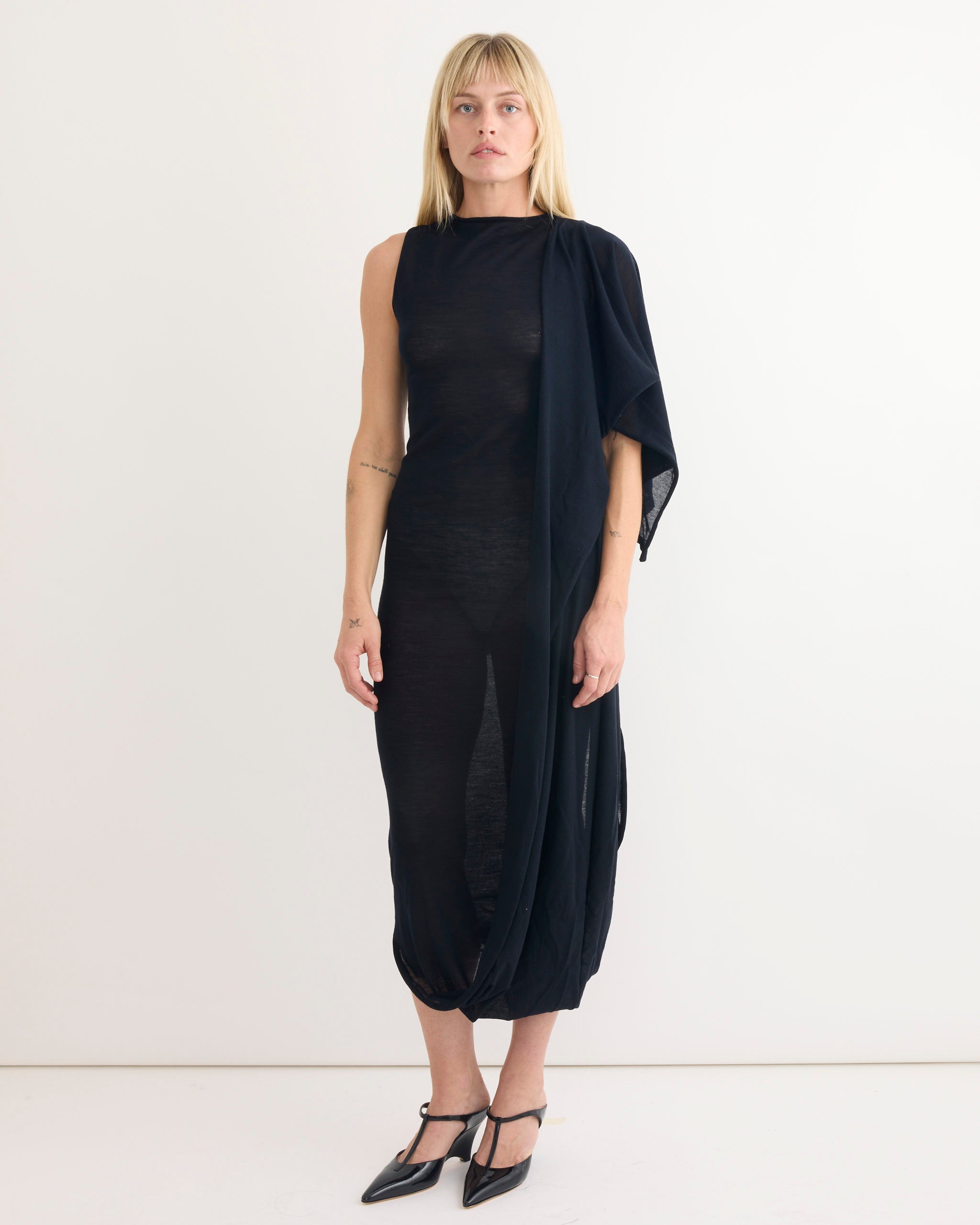 A woman with straight blonde hair and a neutral expression stands against a plain white background wearing the Paloma Wool Willy Dress in Navy—sleeveless, semi-sheer, with an asymmetric draped detail—and black pointed-toe heels.