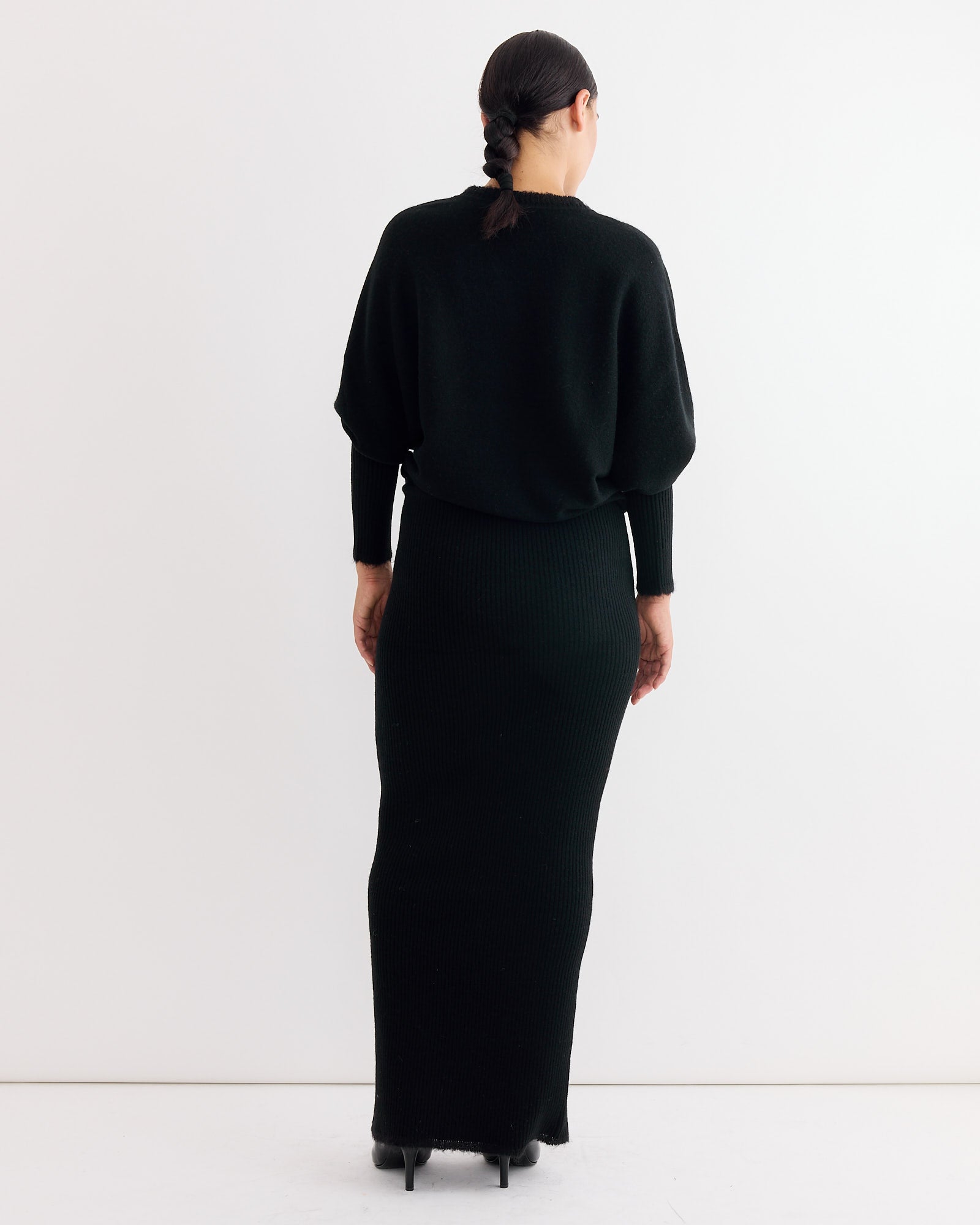 A person with dark hair in a low braid faces away, wearing the By Malene Birger Gislaine Dress in Black—a long-sleeve knit maxi. The background is plain white.