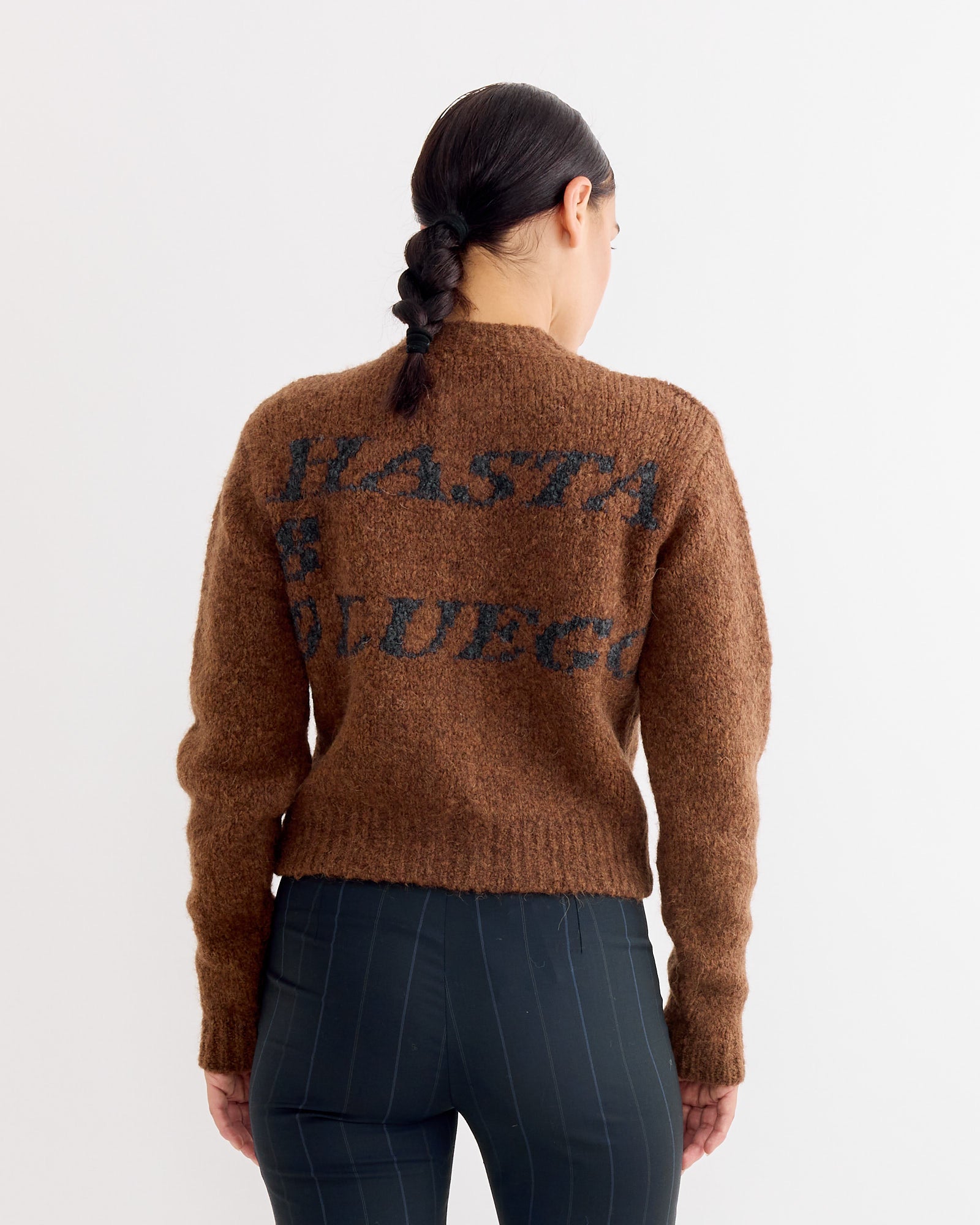A person with dark braided hair, seen from behind, wears the Paloma Wool Hasta Luego Zip in Brown with dark pinstriped pants against a plain white background—ideal for previewing fit and style before your next online chat.