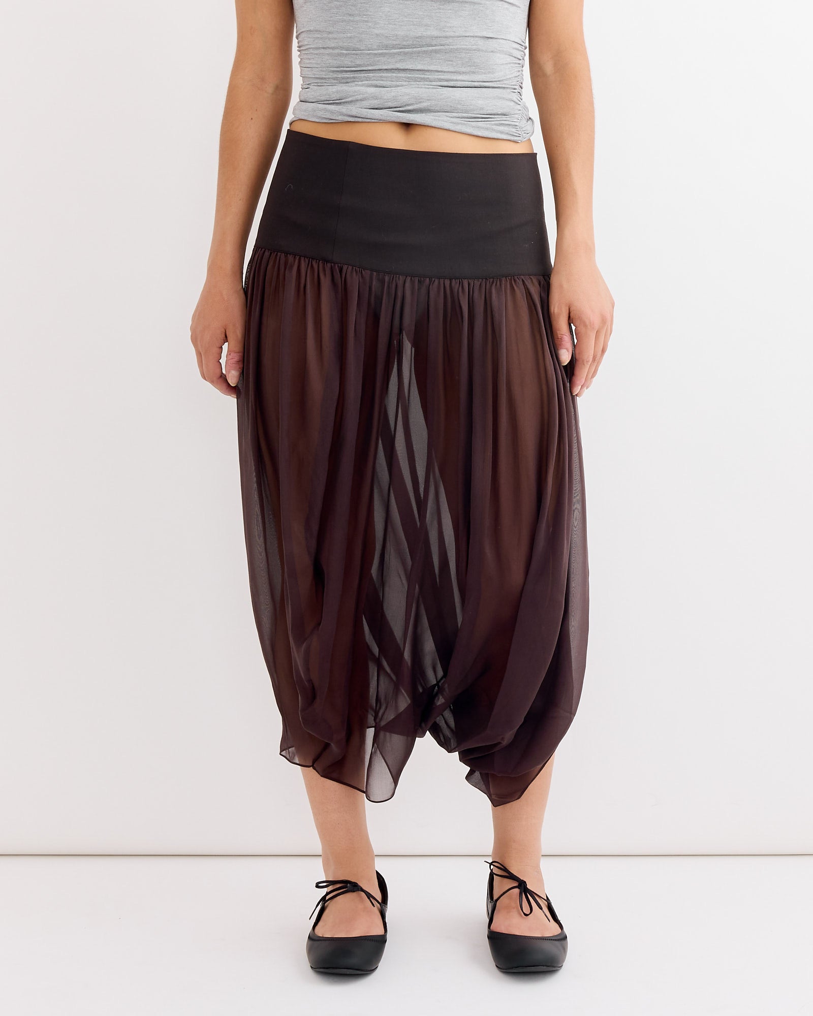 A person wears a gray sleeveless top and the Paloma Wool Mila Skirt in Brown—a sheer silk skirt with a tailored waistband that flows and reveals their legs. Black ballet flats complete the look.