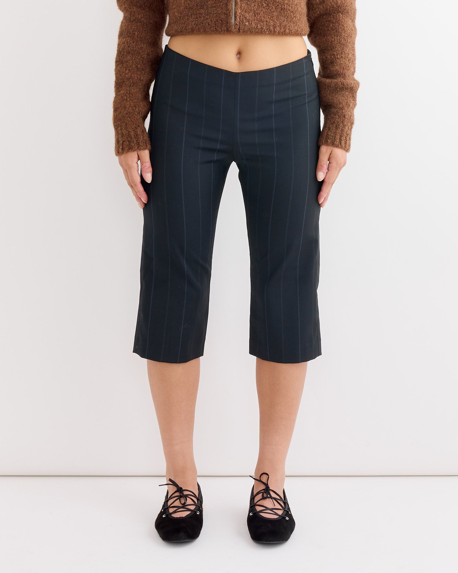 A person wears Paloma Wool's Tinas Pant in Blue—dark, cropped capri pants—paired with a brown long-sleeve sweater and black lace-up shoes, standing against a plain white background. The pants fall below the knees.