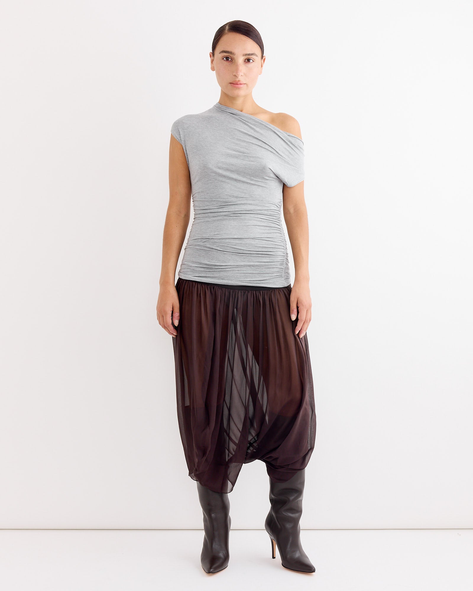 A woman stands against a white background wearing a gray one-shoulder ruched top, Paloma Wool’s Mila Skirt in Brown with a tailored waistband and sheer silk fabric, and knee-high black heeled boots. Her hair is pulled back and she has a neutral expression.