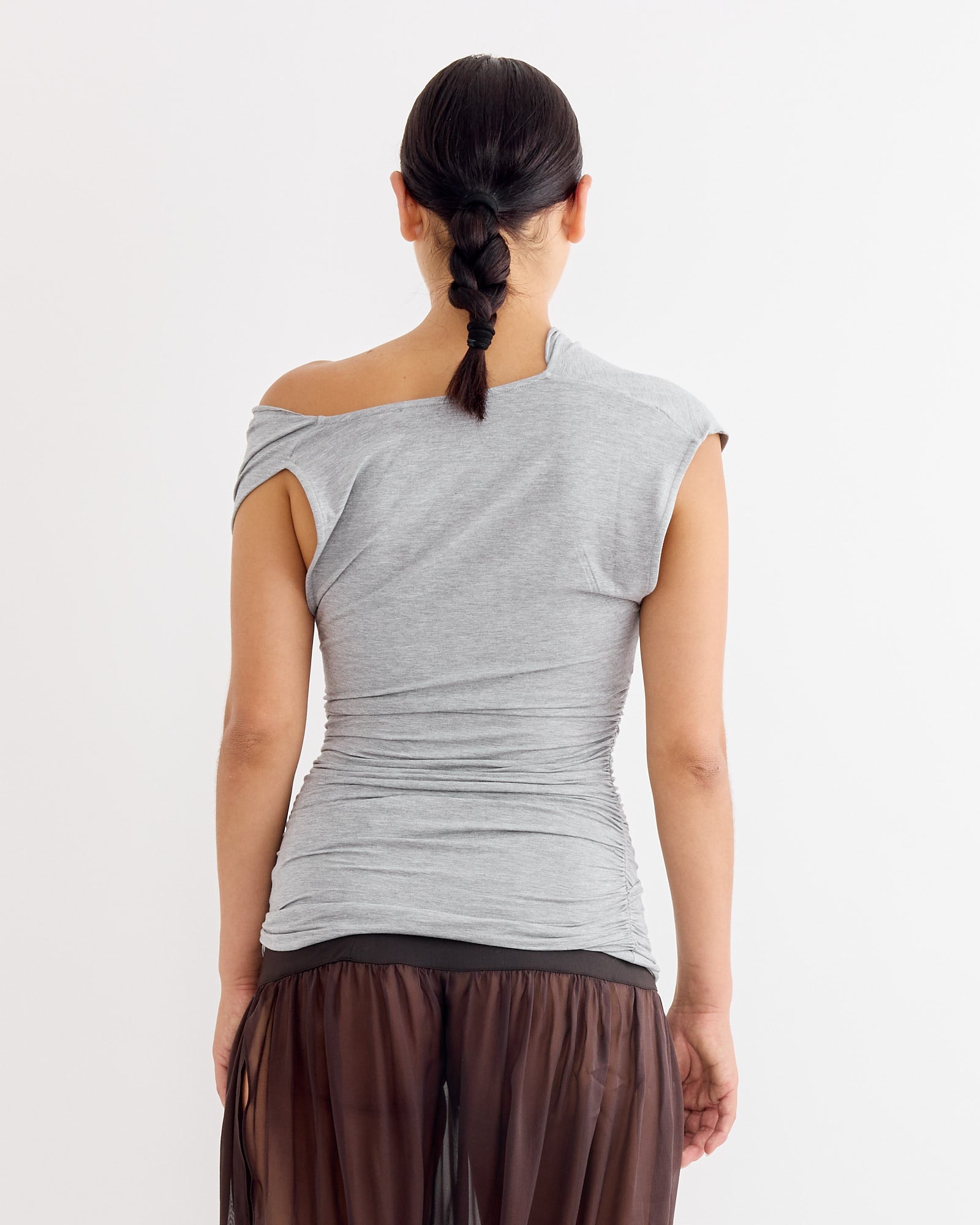 A person with dark braided hair stands facing away, wearing the fitted Paloma Wool Serafin Top in Grey—crafted from stretch micromodal with a draped neckline—and a sheer brown skirt. The background is plain white.