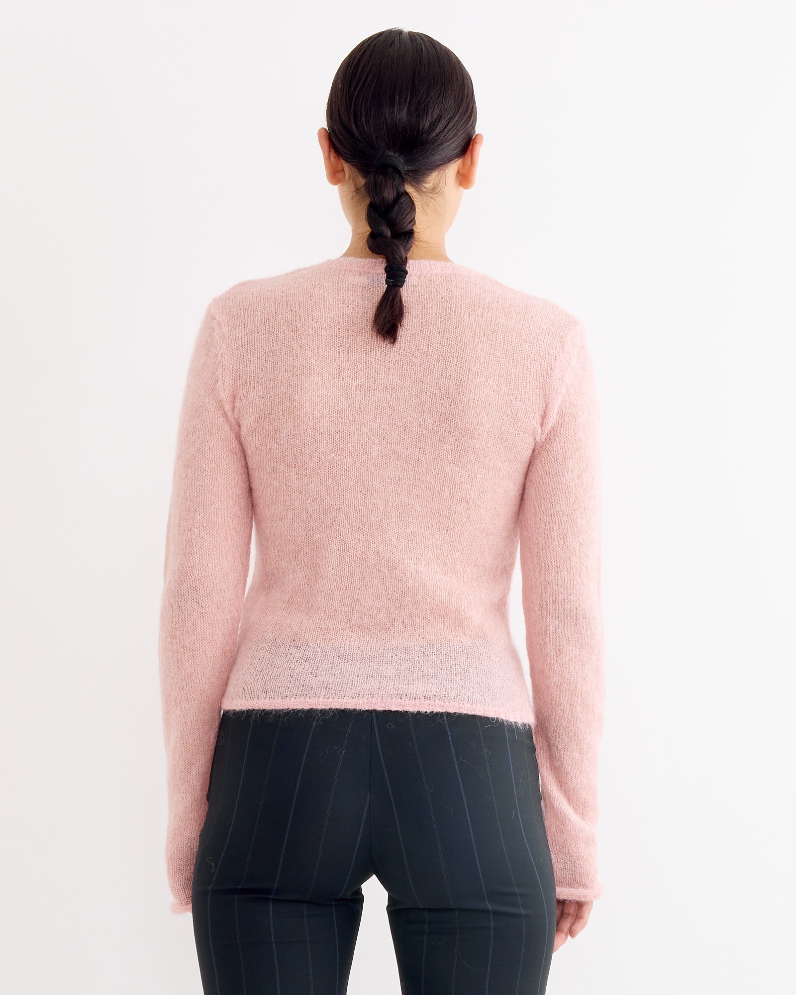 A person with dark braided hair faces away from the camera, wearing the Paloma Wool Tantal Cardigan in Pink and dark pinstriped pants, set against a plain white background.