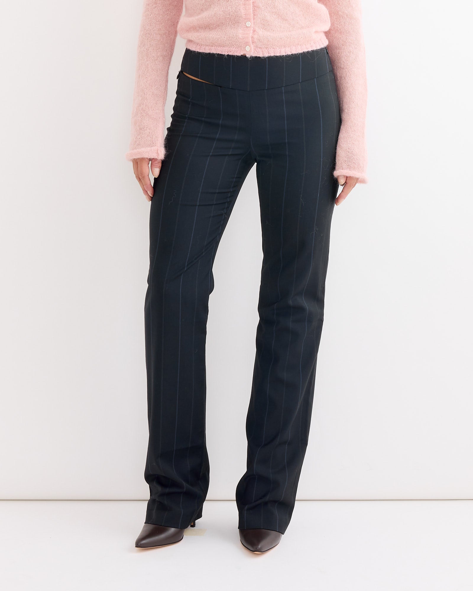 A person wears a light pink long-sleeve top and the Paloma Wool Saba Pant in Blue, featuring high-waisted, straight-leg styling, paired with pointed-toe dark shoes; the pants stand out against a plain white wall.