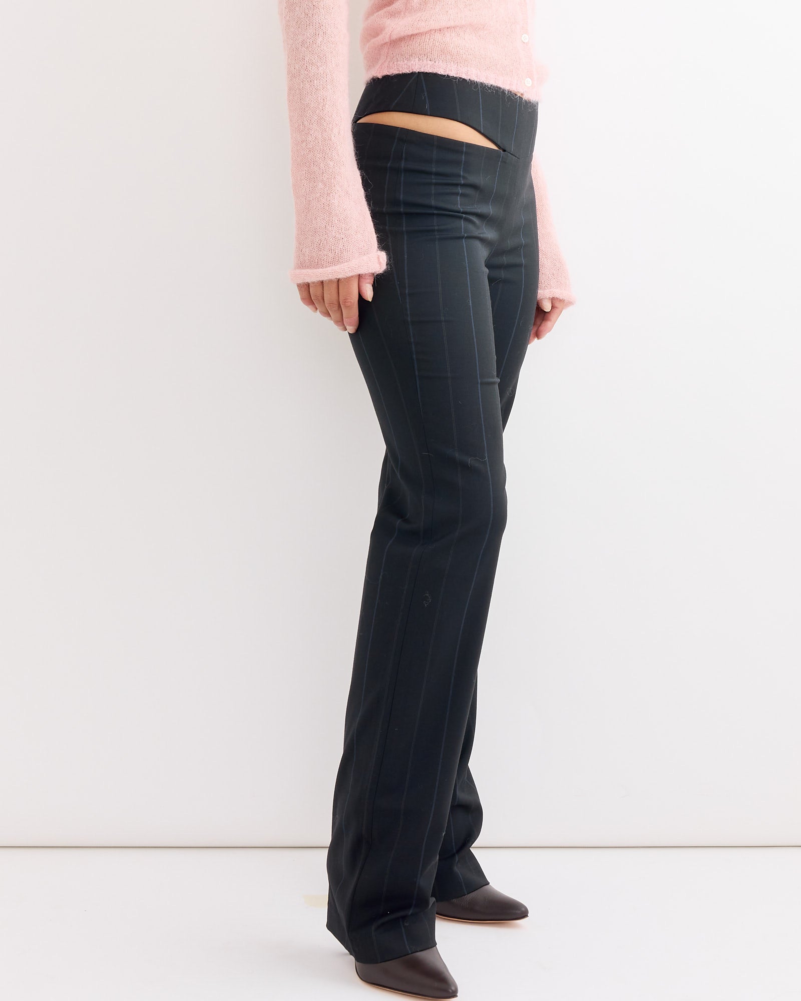 A person wears blue Paloma Wool Saba Pant with a V-shaped waist cutout, a light pink long-sleeve top with flared cuffs, and pointed brown shoes, standing against a plain white background.