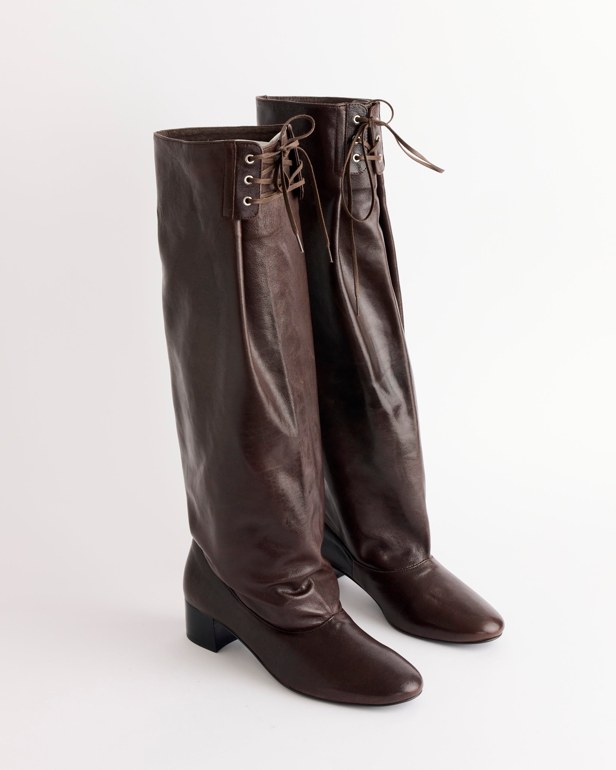 A pair of Paloma Wool Victoria Boot in Brown, tall dark leather boots with low heels and front lace-up detail, stand upright on a white background.