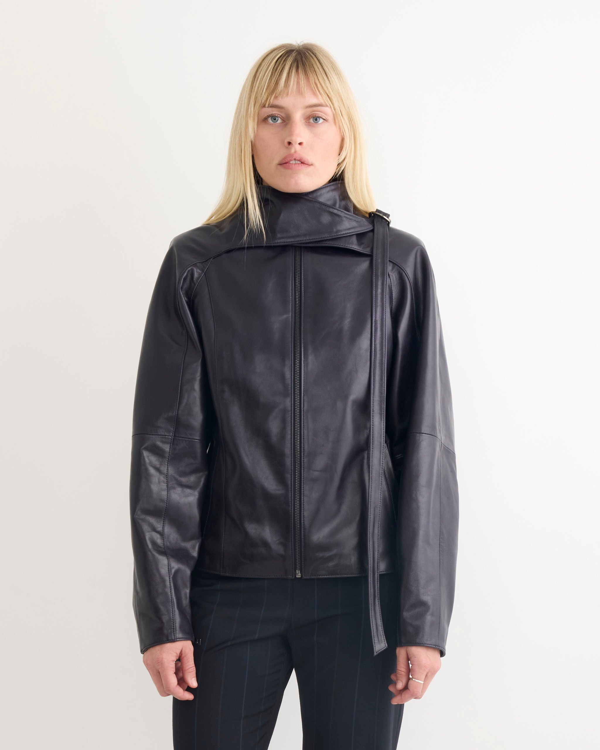 A person with straight blonde hair wears the Paloma Wool Plomy Leather Jacket in Black, which features an asymmetrical collar and long sleeves, standing against a plain white background.