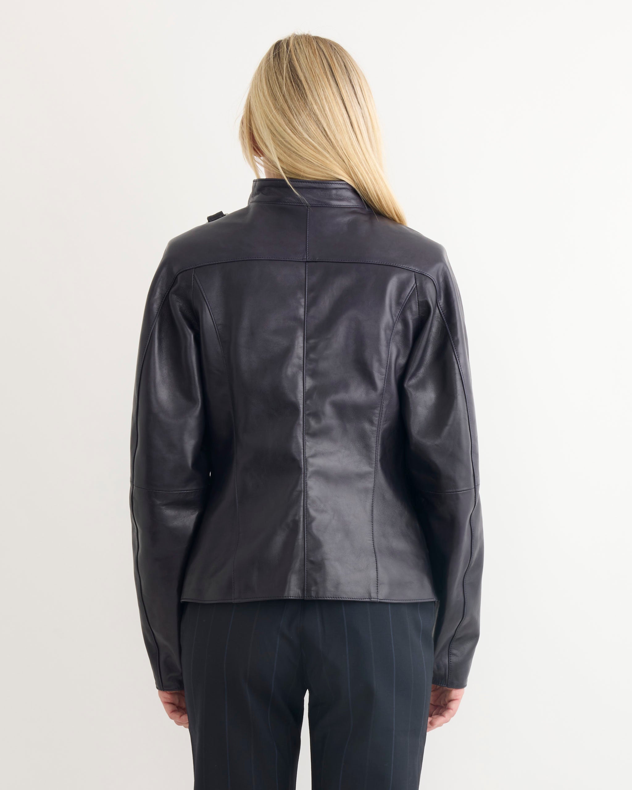 A person with long blonde hair is seen from behind, wearing the Paloma Wool Plomy Leather Jacket in Black and dark pinstriped pants against a plain white background.