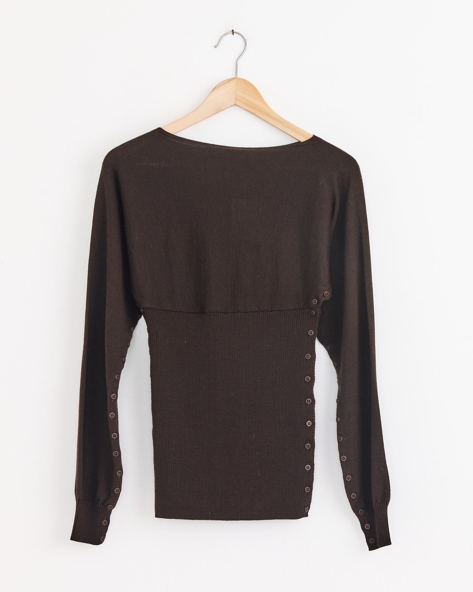 The Paloma Wool Fa Sweater in Brown, crafted in Spain from extrafine merino wool, has a wide neckline and side button details. Shown hanging on wood against a white background, this long-sleeve piece blends style and comfort.