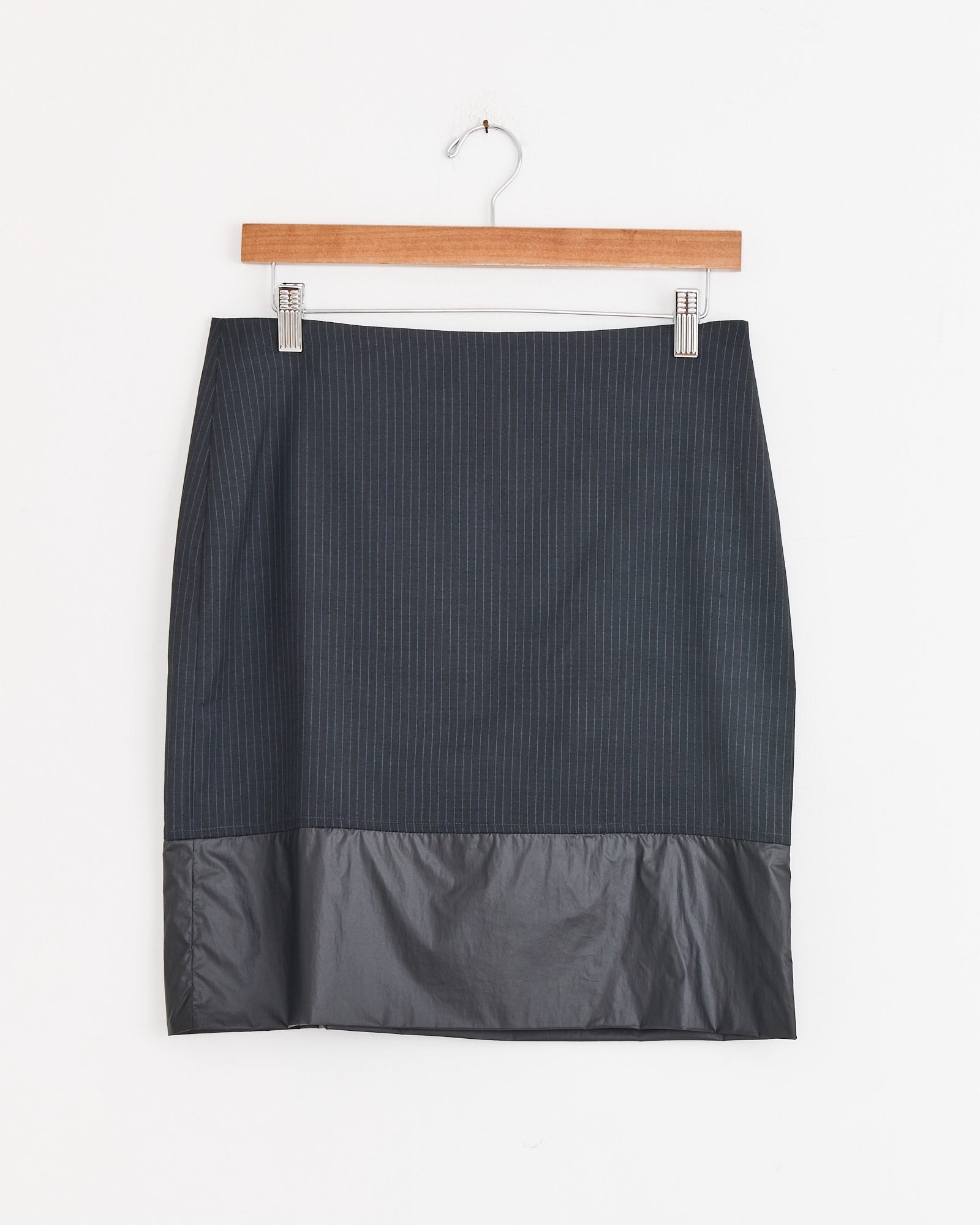 The Ongi Skirt in Grey by Paloma Wool is a knee-length suiting skirt with subtle pinstripes and a shiny solid hem, shown on a wooden hanger against a white background.