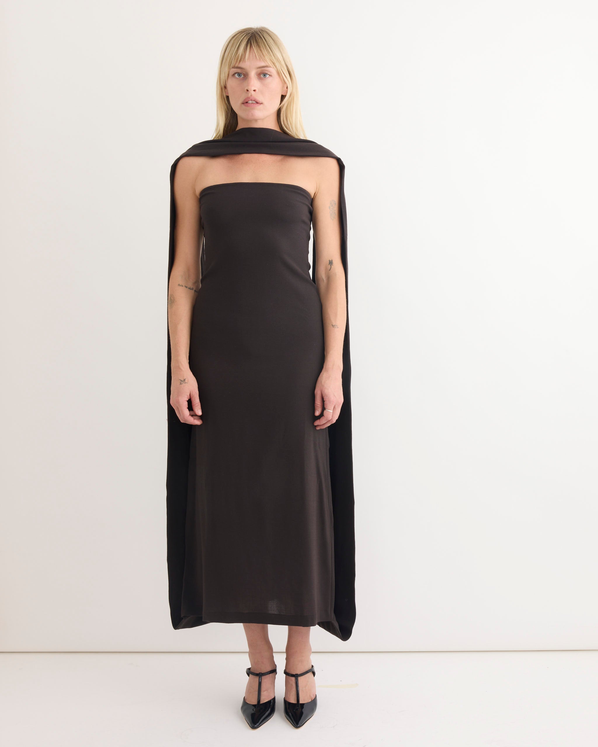 A woman with blonde hair and bangs stands against a white background, wearing the strapless Paloma Wool Alberch Dress in Dark Grey with a matching draped cape and black pointed heels, her expression neutral.