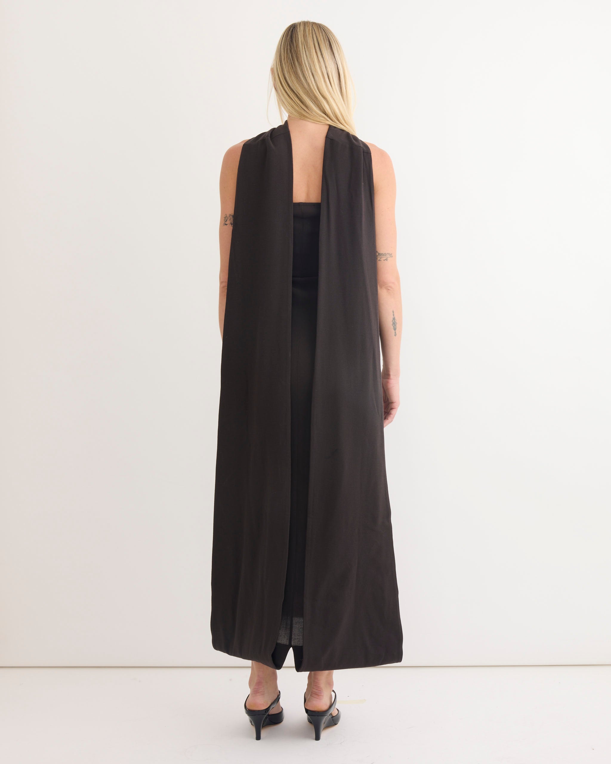 A person with long blonde hair, wearing the Paloma Wool Alberch Dress in Dark Grey—a sleeveless, floor-length wool blend dress with an open back—stands facing a white wall in a minimal, well-lit setting. Black heeled sandals are visible.