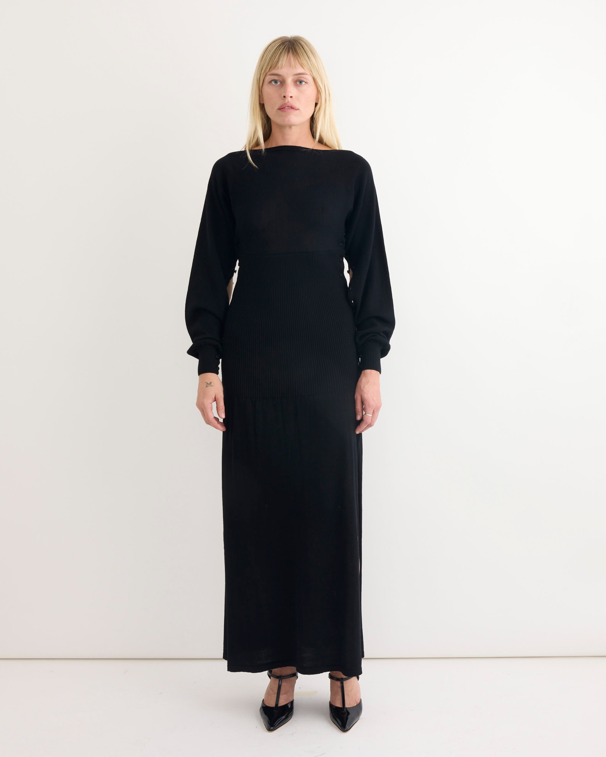 A person with blonde hair wears the Paloma Wool Fa Dress in Black with black heels, standing facing forward against a plain white background.