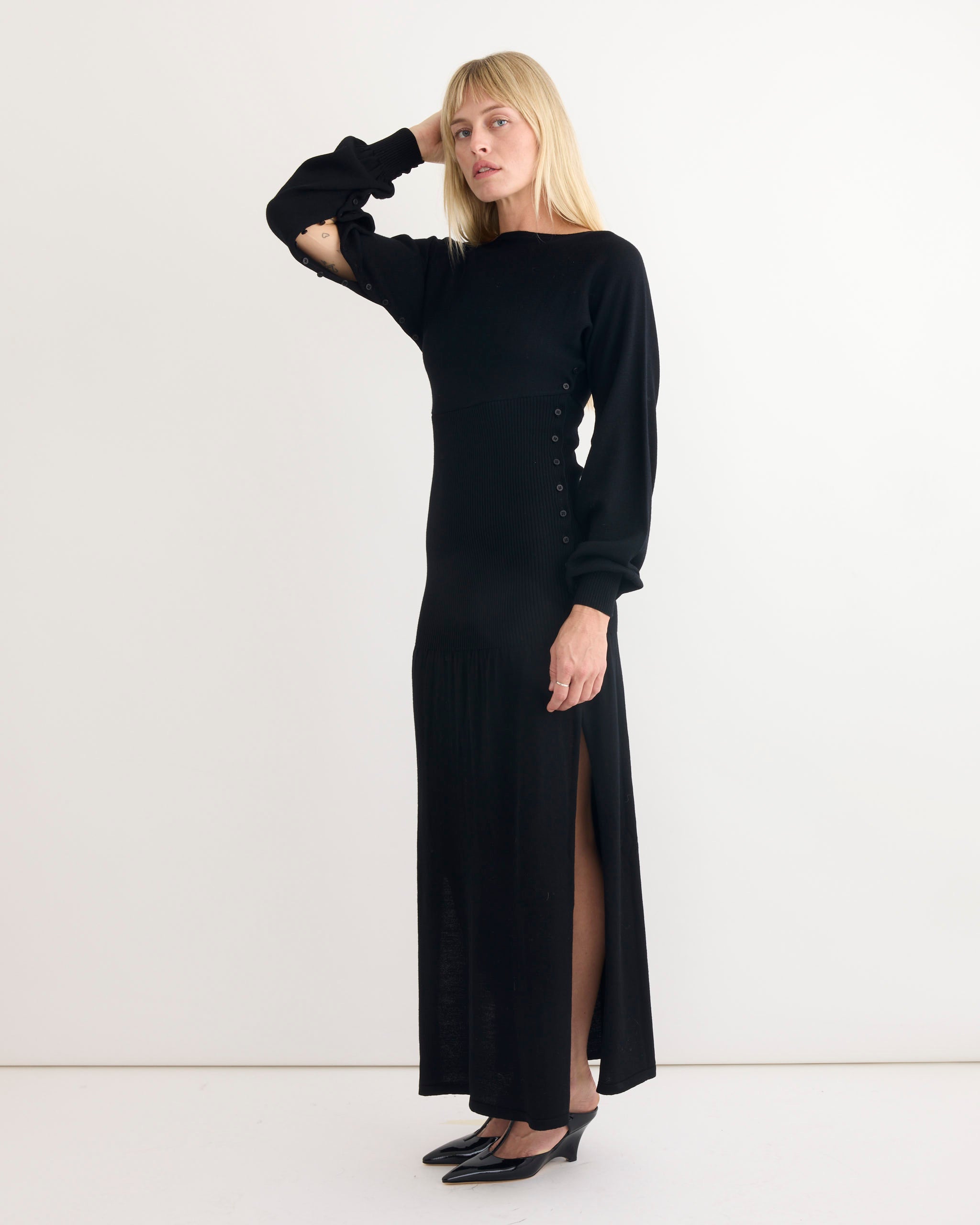 A woman wears the Paloma Wool Fa Dress in Black—a long ribbed dress with side buttons and a high slit—paired with black heeled shoes. She stands against a white background, touching her hair and looking to the side.