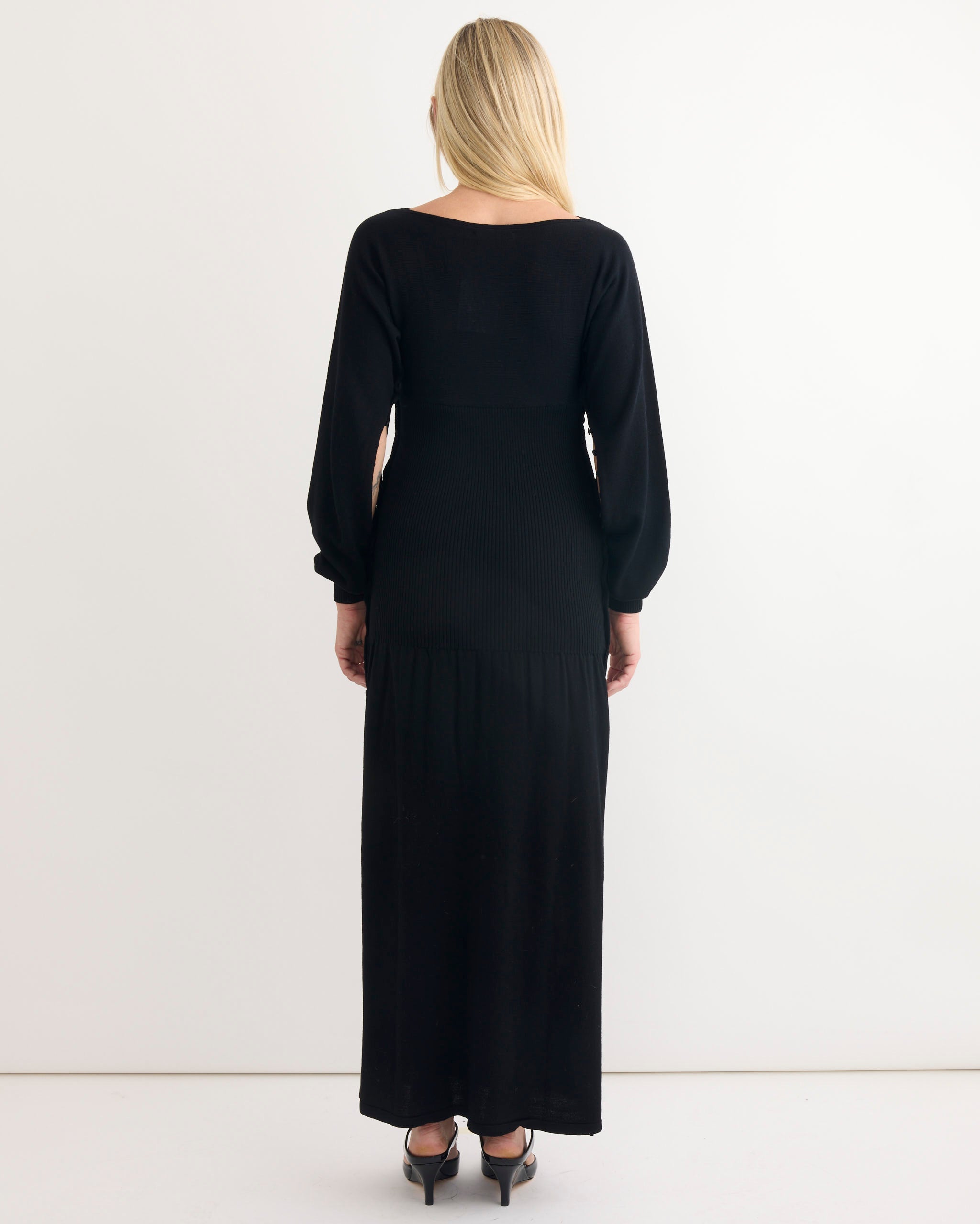 A woman with long blonde hair faces away, wearing the Paloma Wool Fa Dress in Black—an ankle-length, long-sleeved ribbed dress—paired with black heeled shoes against a plain white background.