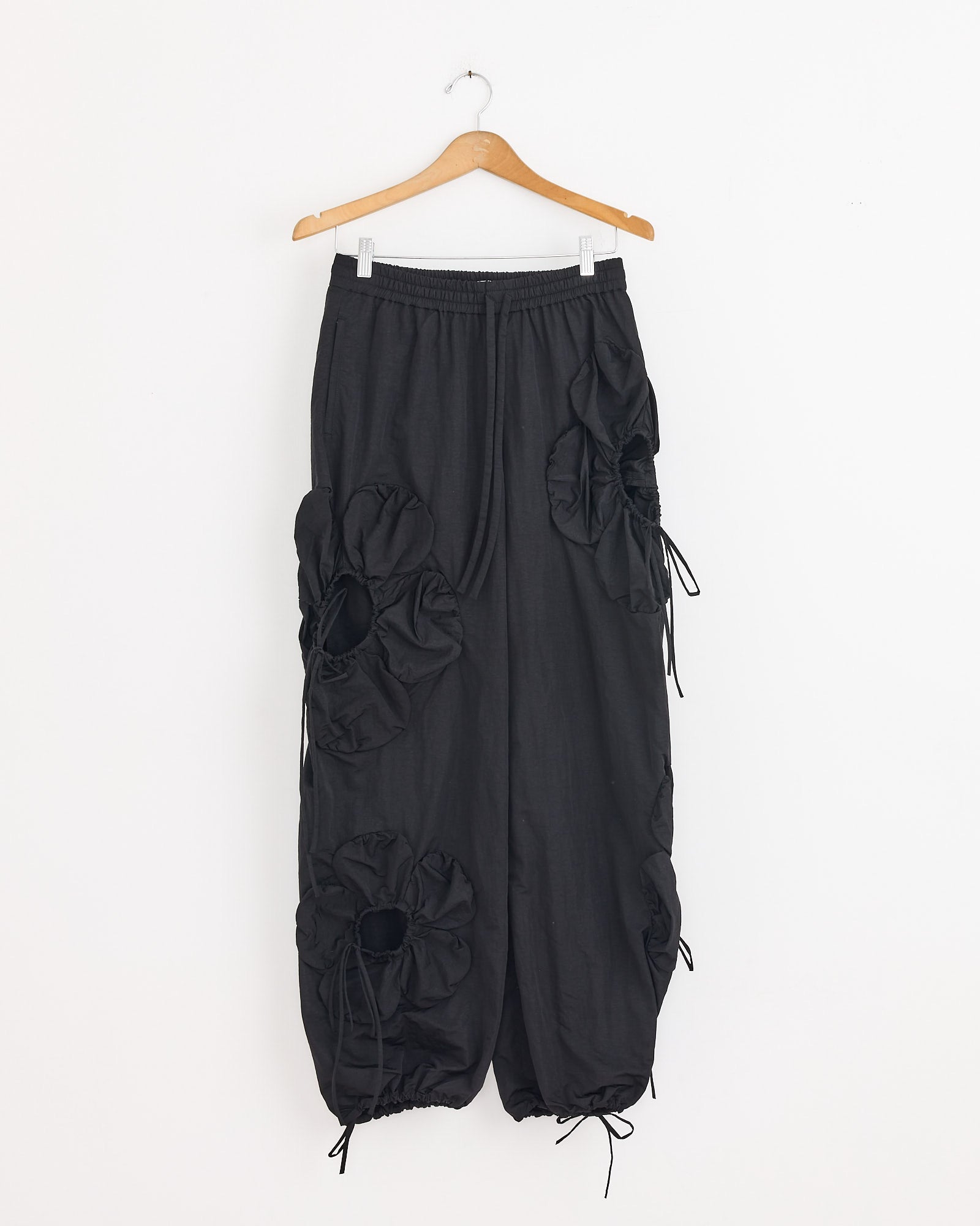 The Flower Pant in Black by J. Kim features a baggy silhouette, elastic waistband, sculptural flower details, and drawstrings, creating a unique statement piece. Shown hanging on a wooden hanger.