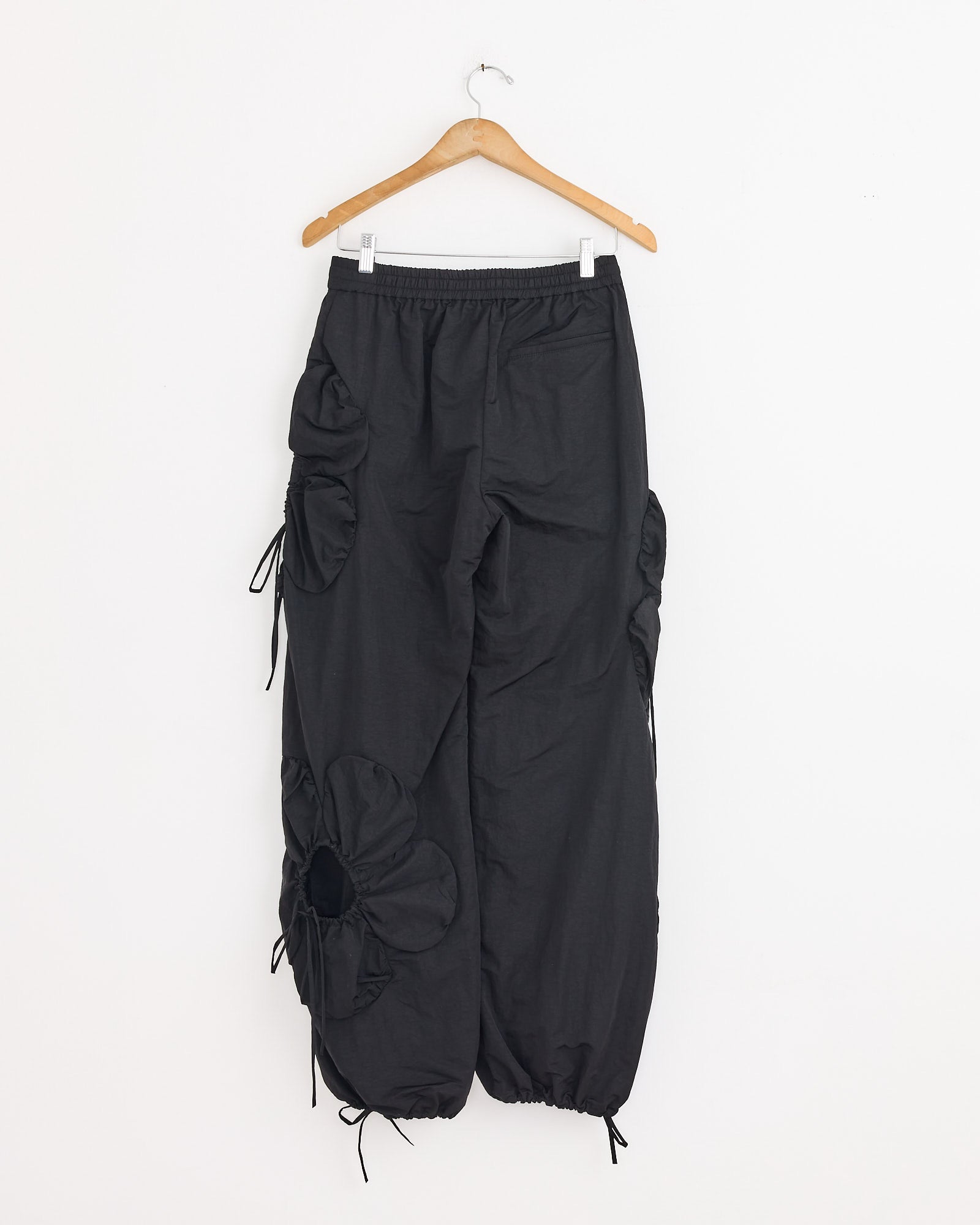 The J. Kim Flower Pant in Black features a loose fit, elastic waist and ankles, sculptural flower details with side cutouts on one leg, creating a voluminous silhouette. Shown on a wooden hanger against a white backdrop.