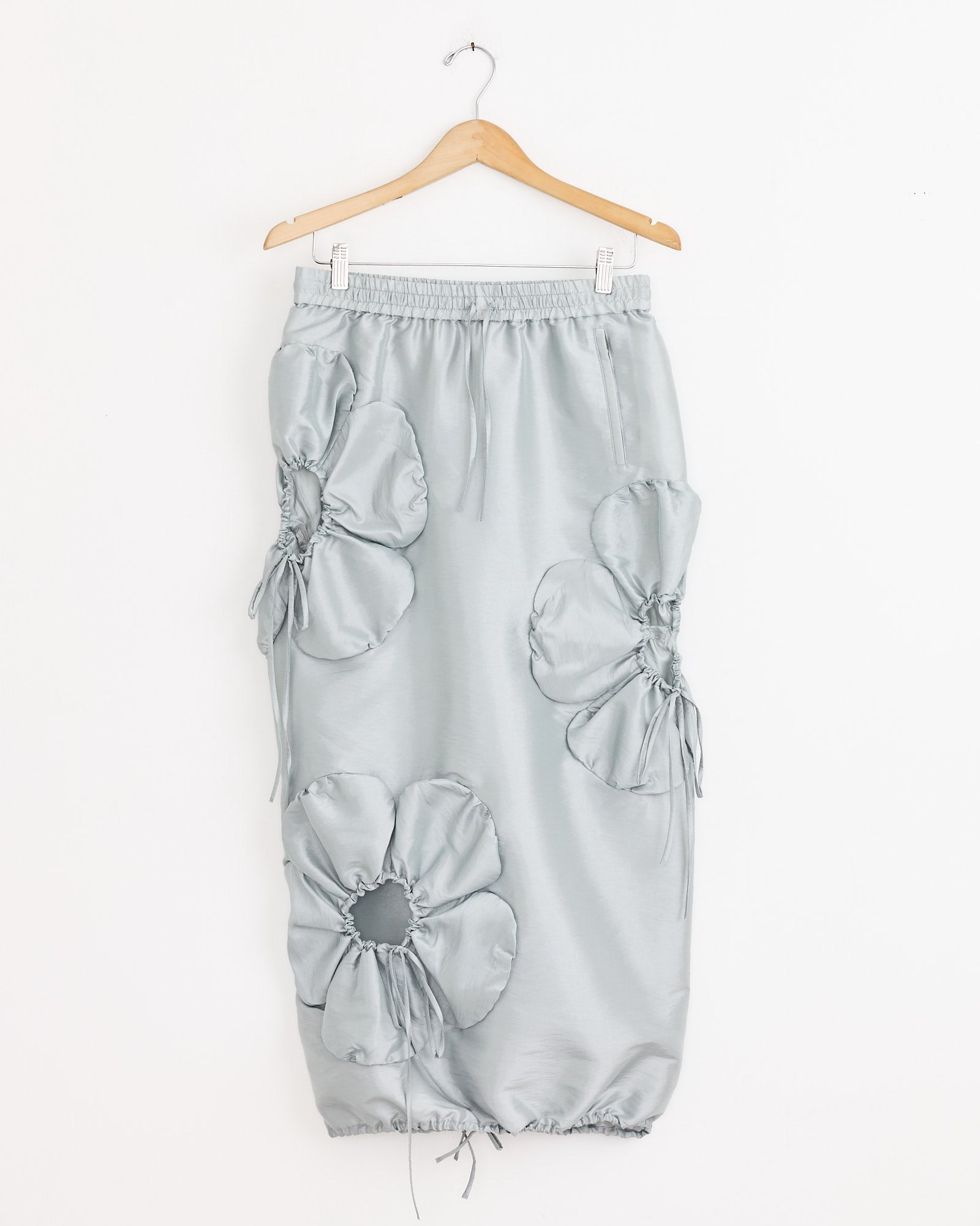 The J. Kim Flower Skirt in Silver is made from lightweight nylon, featuring large gathered adjustable flower cut-outs, ruching details, an elastic waistband, and a drawstring hem. It hangs on a wooden hanger.