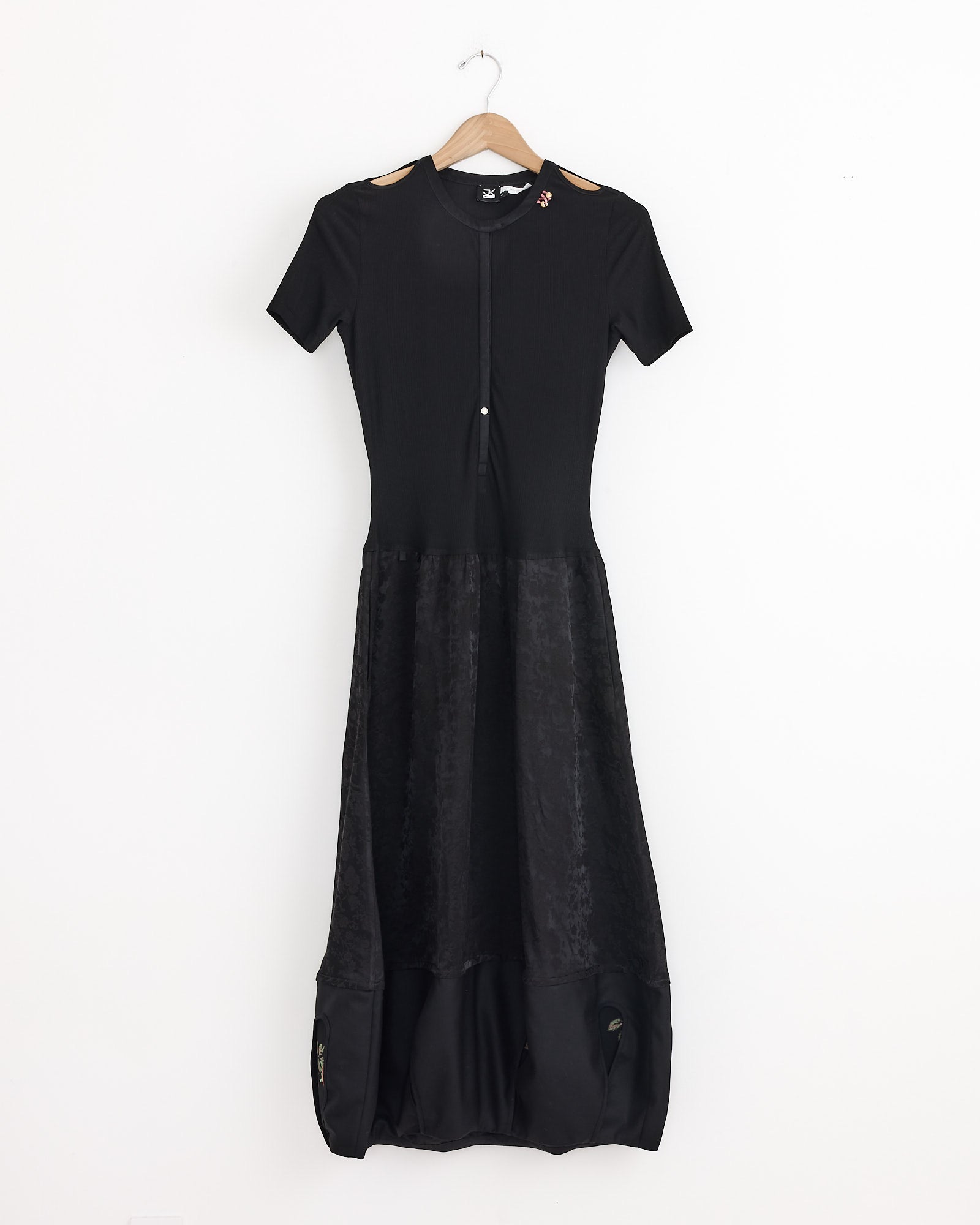 The J. Kim Rattle Dress in Black, featuring short sleeves, a fitted top, jacquard viscose skirt, and button detail at the neckline, hangs on a wooden hanger against a plain white background.