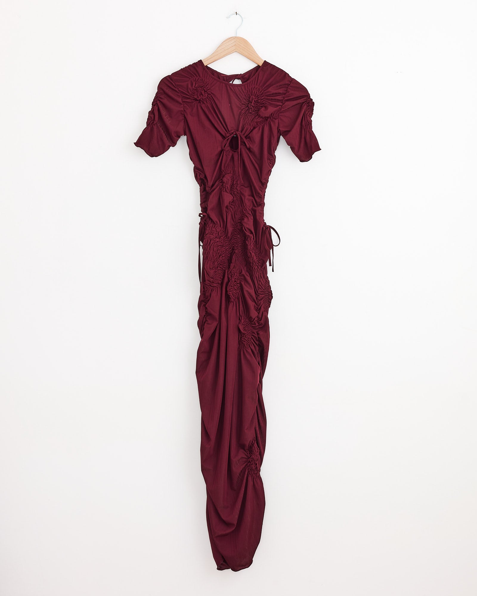 The J. Kim Markiza Dress in Burgundy, featuring short sleeves, gathered and ruched details, and a chest keyhole cutout, hangs on a wooden hanger against a white wall.