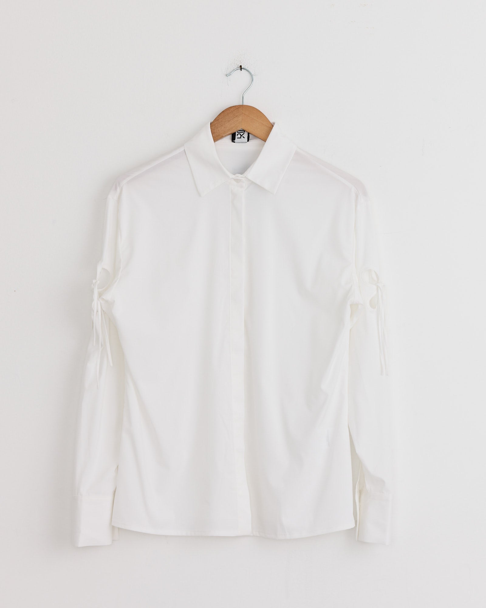 The J. Kim Petal Shirt in White, a crisp cotton poplin long-sleeve with collar and sleeve ties at the elbows, hangs on a wooden hanger against a plain white wall—an elegant update to a classic piece.