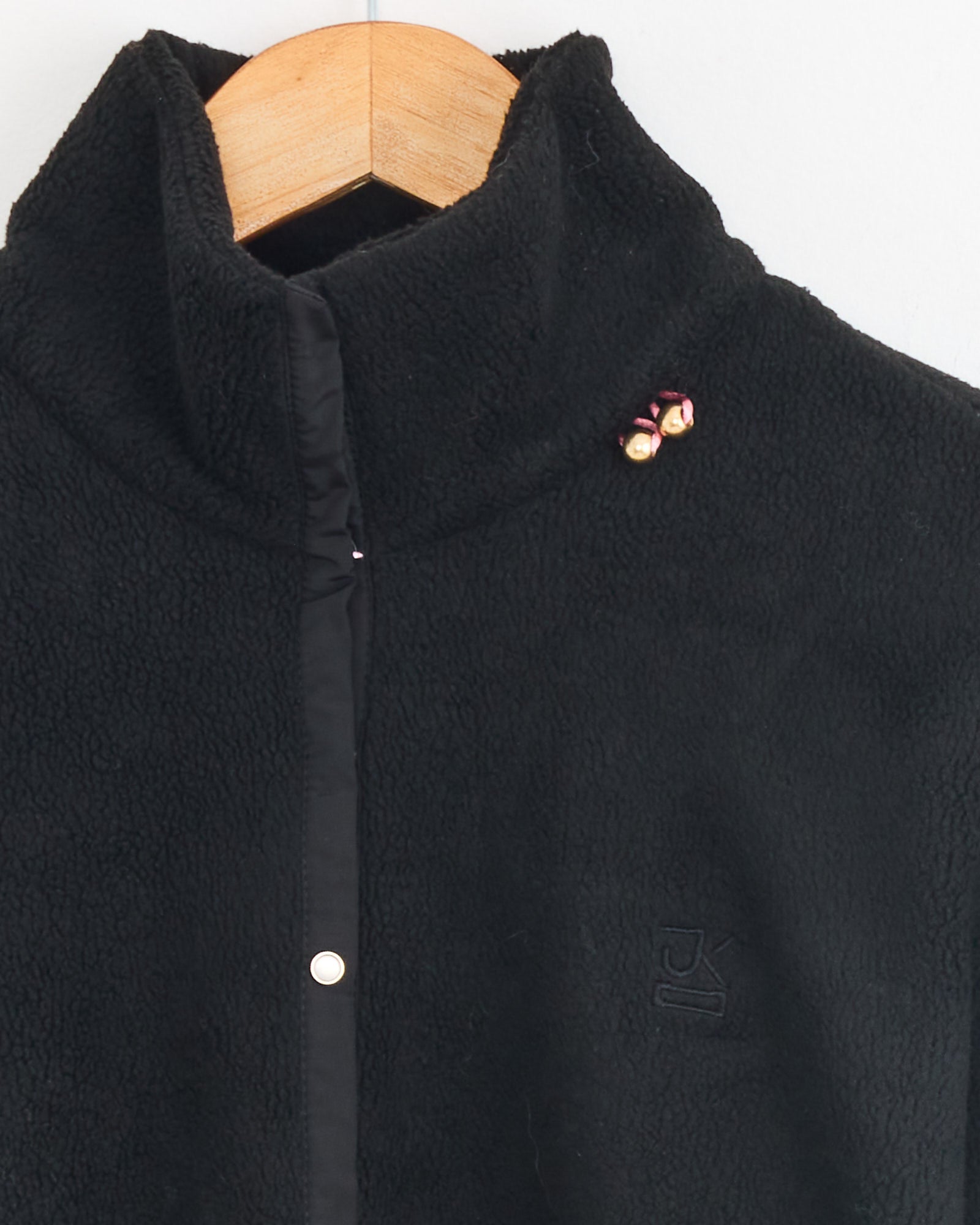 The J. Kim Bellflower Fleece Sweater in Black hangs on a wooden hanger, featuring a high collar, buttoned front, subtle embroidered chest logo, and two small pink and yellow flower pins near the collar.