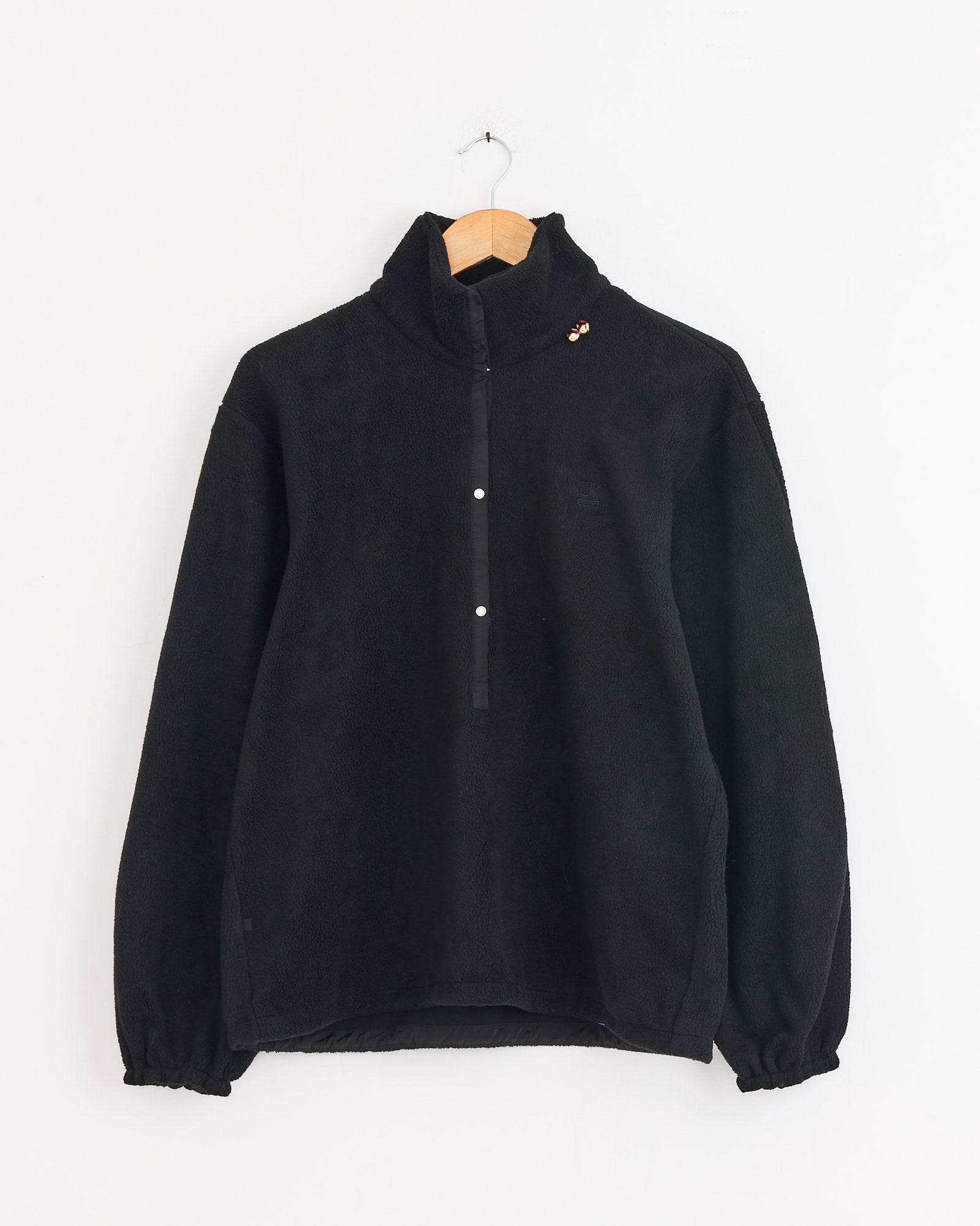 The J. Kim Bellflower Fleece Sweater in Black is a vintage-inspired pullover with a half-zip front, snap buttons, elastic-cuffed long sleeves, and a small pin on the left shoulder. Shown on a wooden hanger against a white wall.