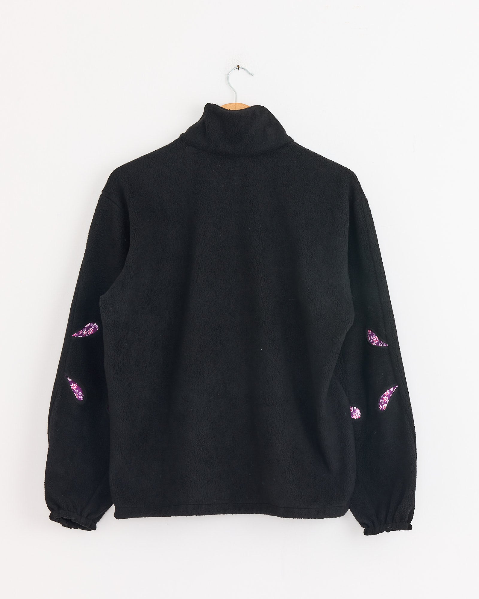 The J. Kim Bellflower Fleece Sweater in Black hangs on a wall, back view. It features purple and white embroidered eye designs on the sleeves, a high collar, and elastic cuffs.