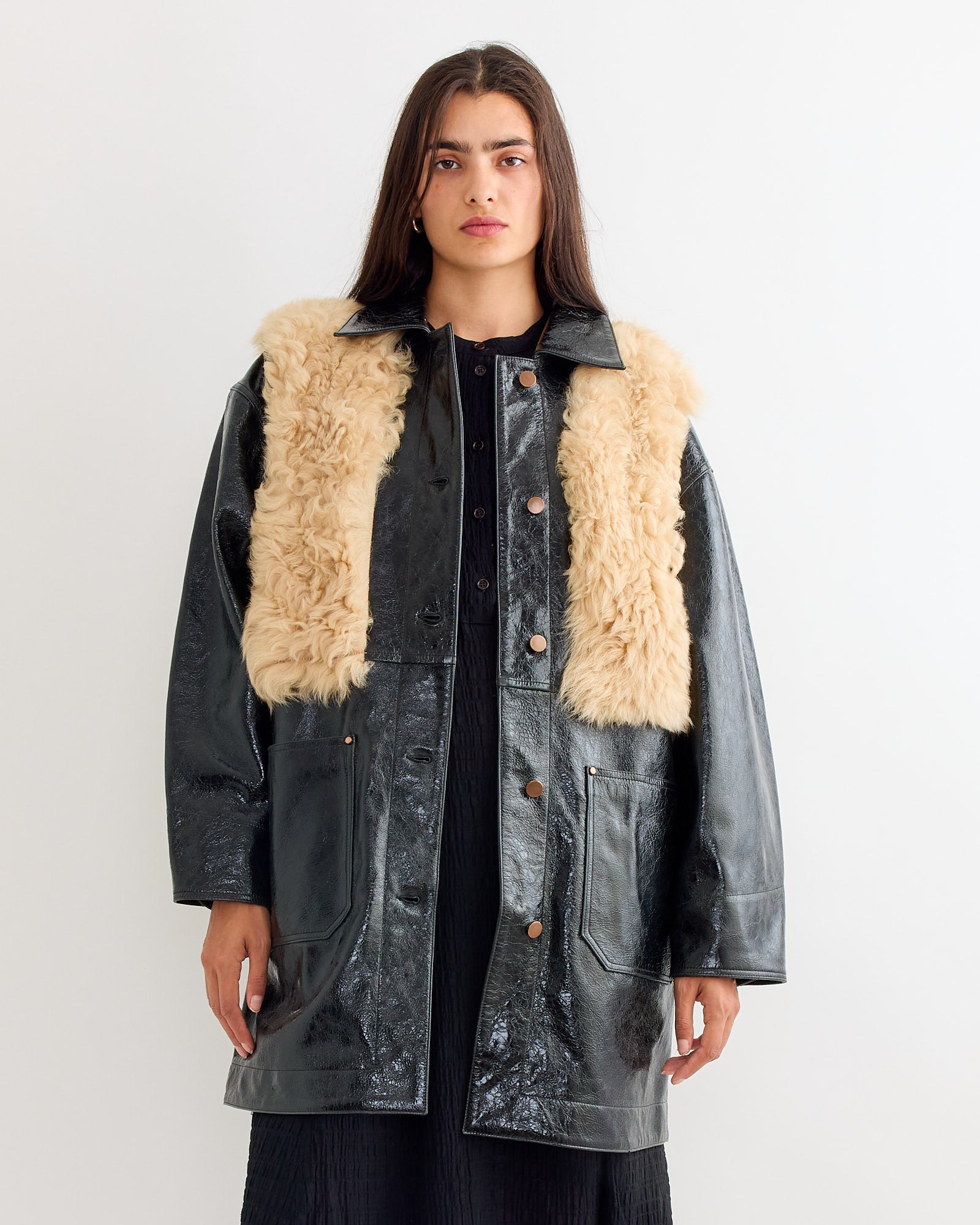 A woman stands facing forward, wearing the Milena Silvano Chore Coat in Black Mix—a shiny black leather jacket with beige faux fur accents—over a black dress, set against a plain light background.